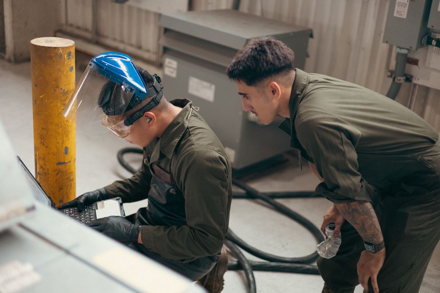 Two Marines in olive uniforms working together on industrial equipment in military workshop.
