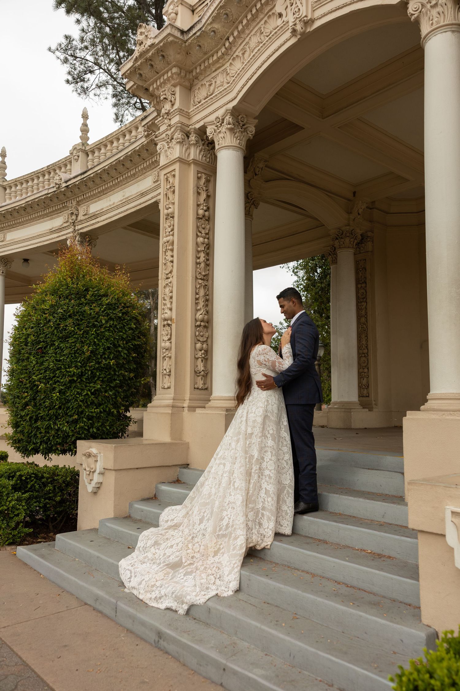 Bride and groom embracing on ornate stone steps of a classical building with tall columns.