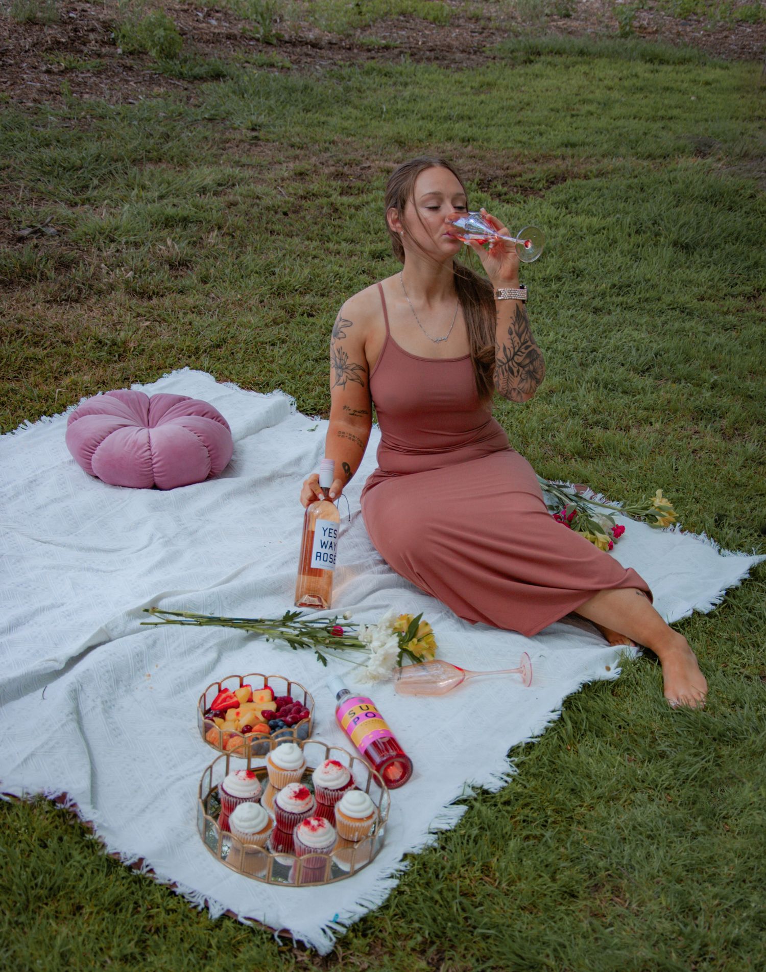 Woman in mauve dress enjoying picnic on white blanket with cupcakes and rosé wine outdoors.