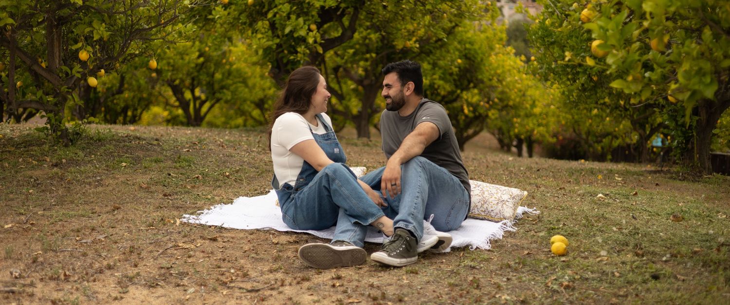 A couple sits together on a blanket in a lush citrus orchard, smiling and holding hands.
