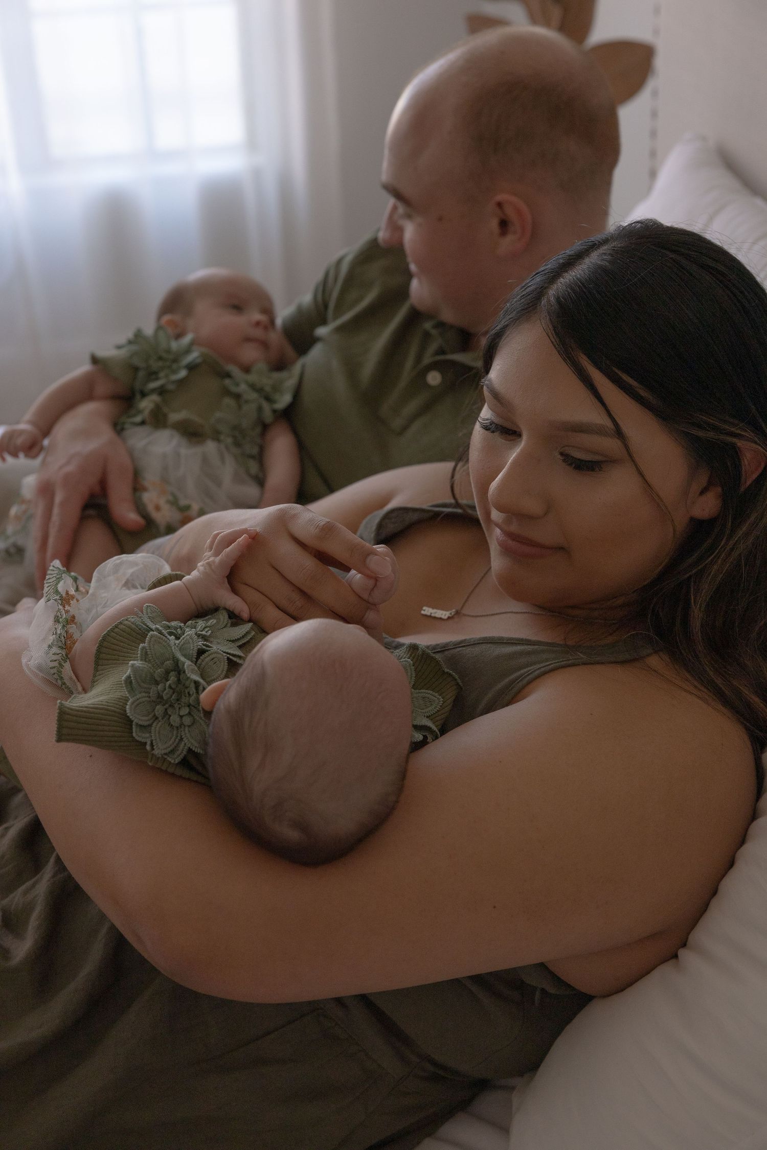 Parents in olive green hold their newborn twins on a cozy bed in a soft, natural light newborn family photo session.