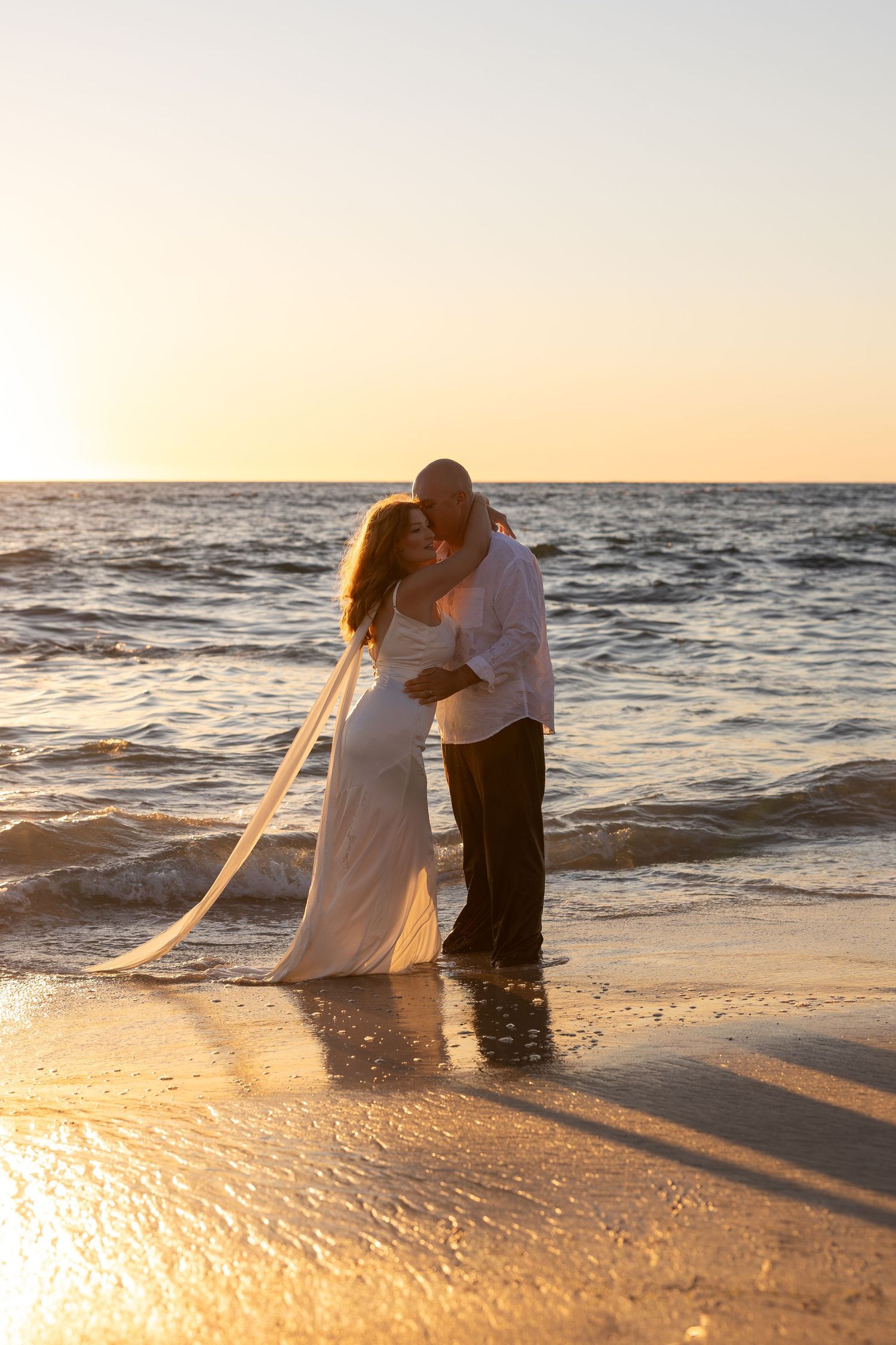 Bride and groom kissing on a sandy beach at golden sunset, waves gently lapping at their feet, veil flowing in the breeze.