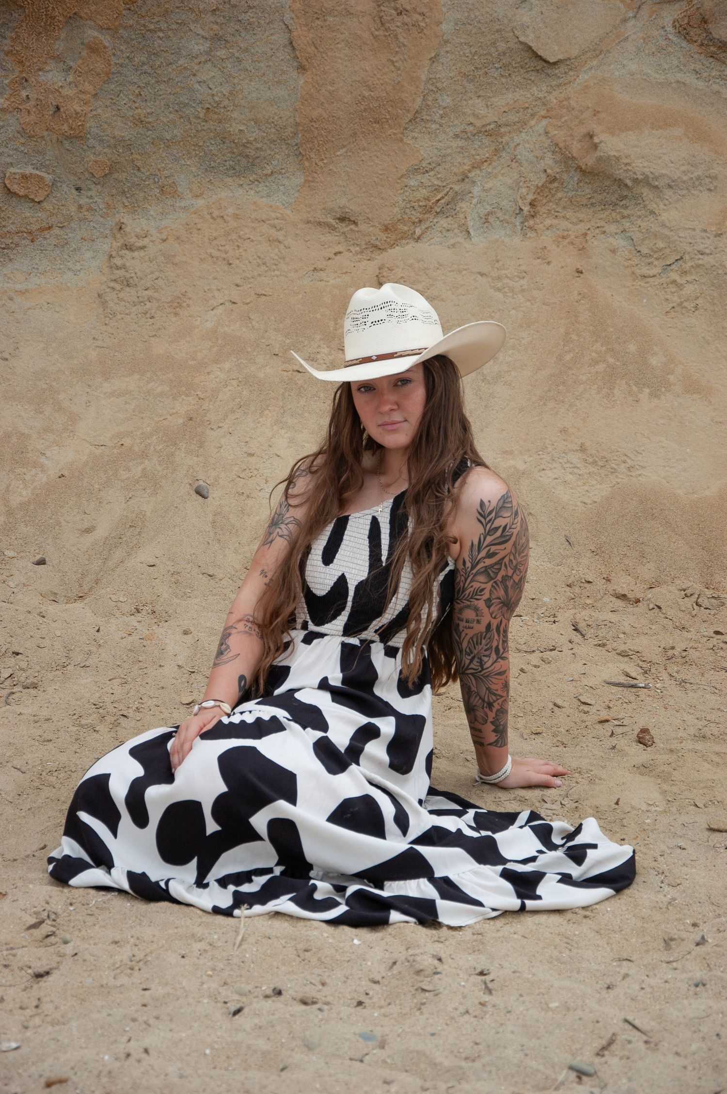 Tattooed woman in black and white maxi dress and cowboy hat sitting on sand.