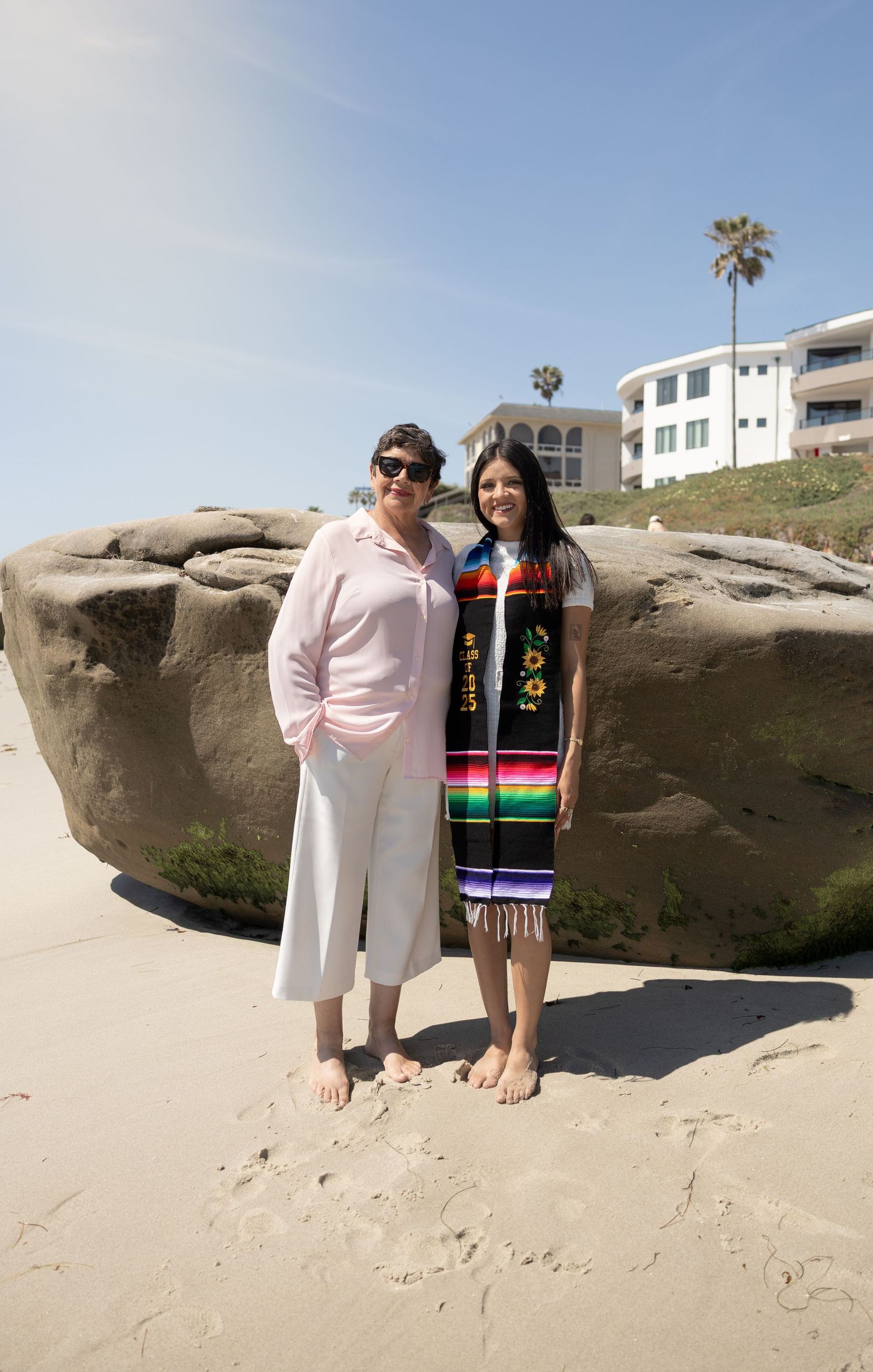 Two women stand barefoot on a sandy beach near large rocks on a sunny day.