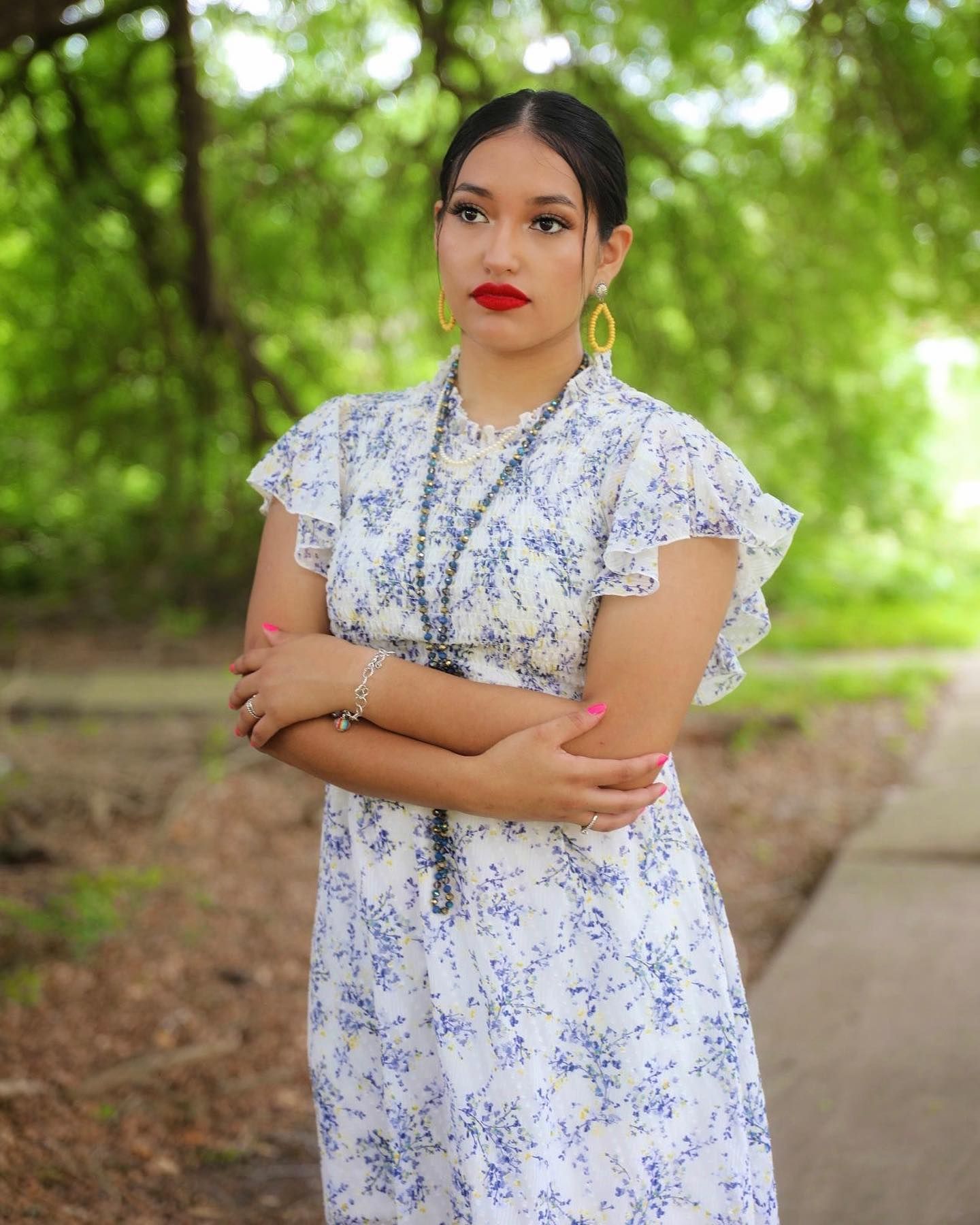 Young woman in blue floral dress with red lipstick poses confidently outdoors among lush green trees.