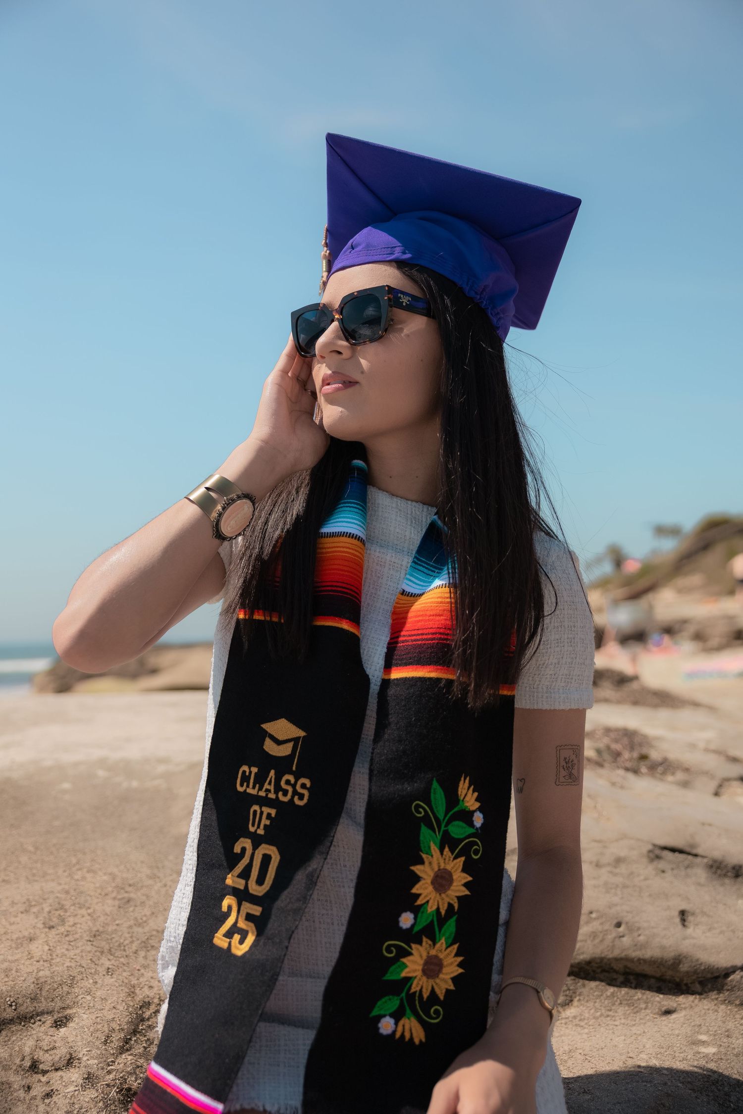 A graduate in a blue cap, sunglasses, and Class of 2025 stole gazes sideways on a sunny beach.