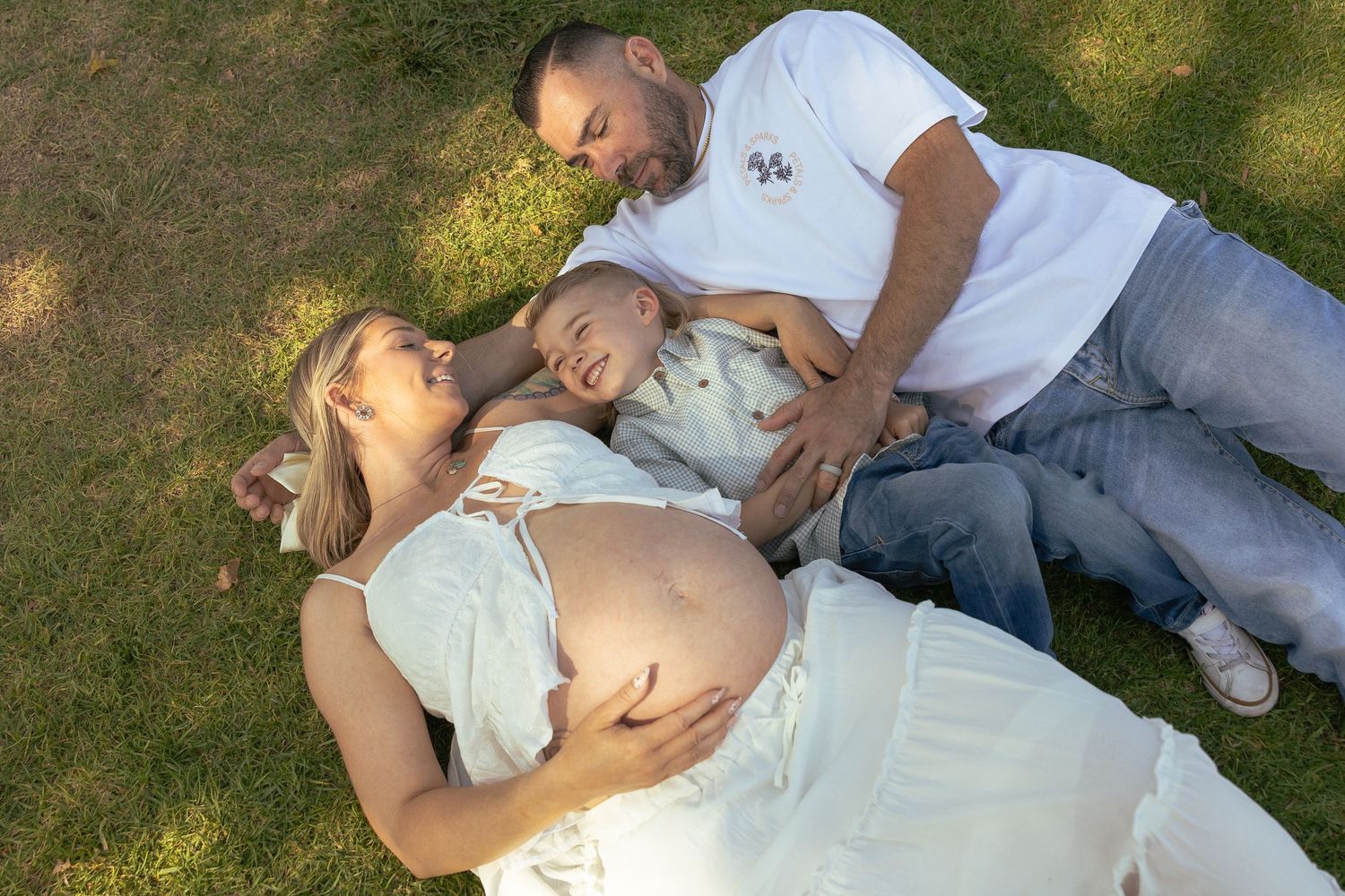 Pregnant woman lying on grass with partner and baby, enjoying a family maternity photo session outdoors.