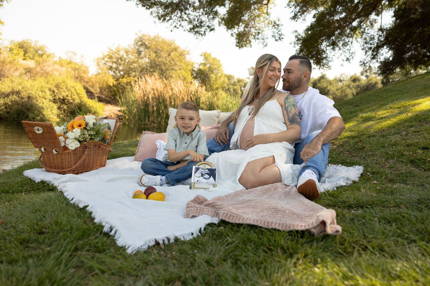Pregnant woman with husband and toddler son enjoying a picnic on grass near a pond during a maternity photo shoot.