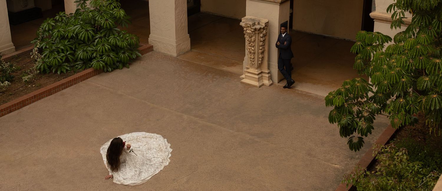 Bride in white wedding dress kneeling in a sunlit courtyard with stone columns and lush greenery.