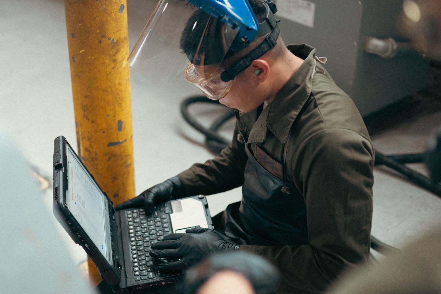 Worker in protective face shield and gloves using rugged laptop in industrial facility.