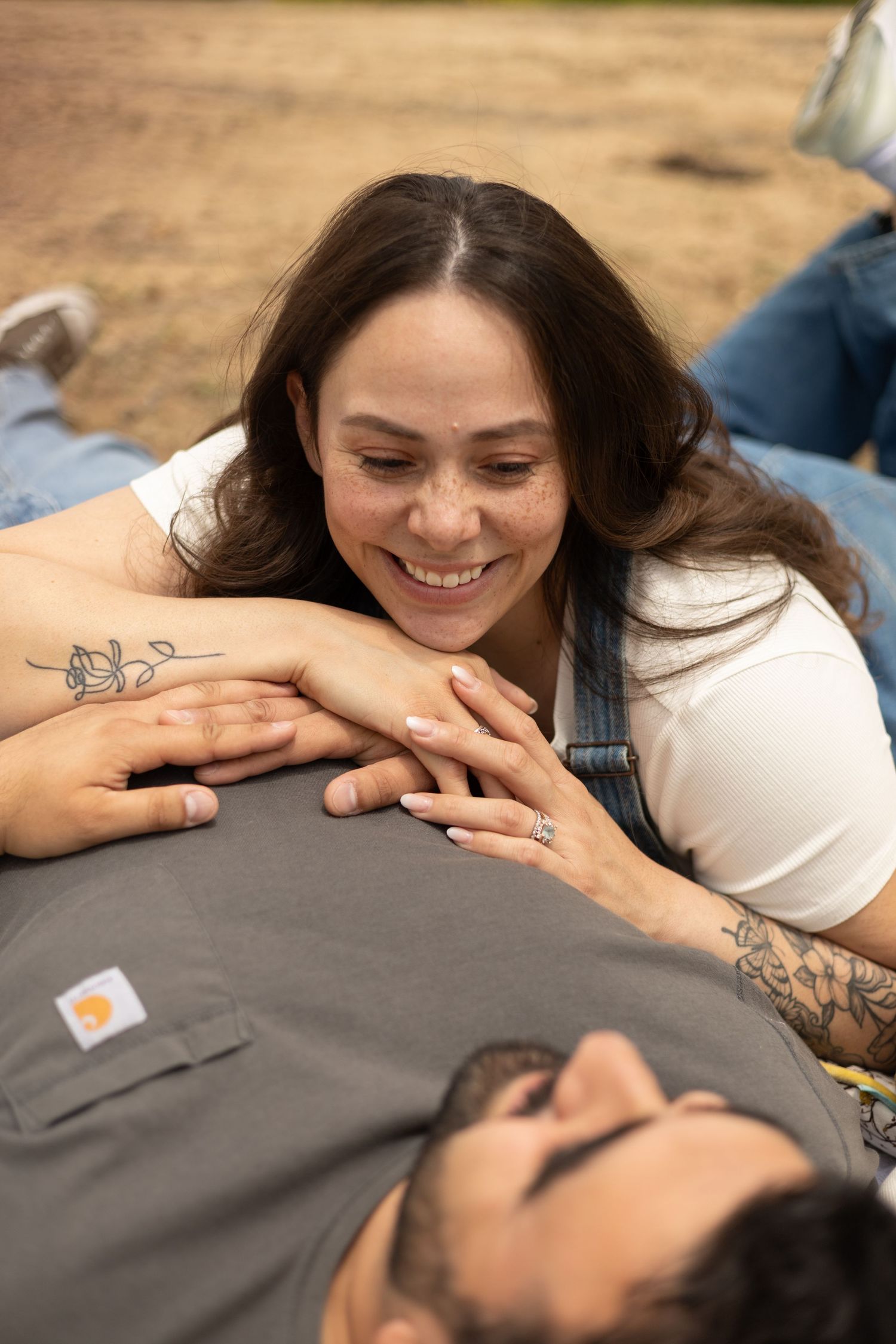 A smiling tattooed woman in overalls rests her head on a mans chest outdoors.