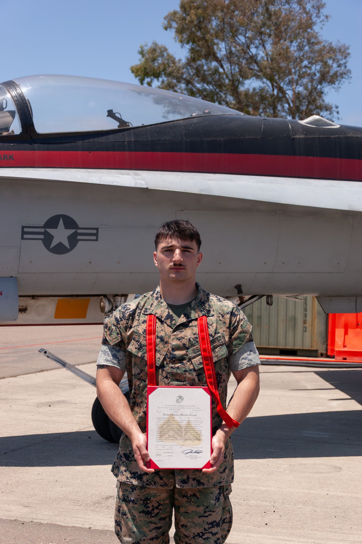 Marine in camouflage uniform holds certificate in front of military jet aircraft on airfield.