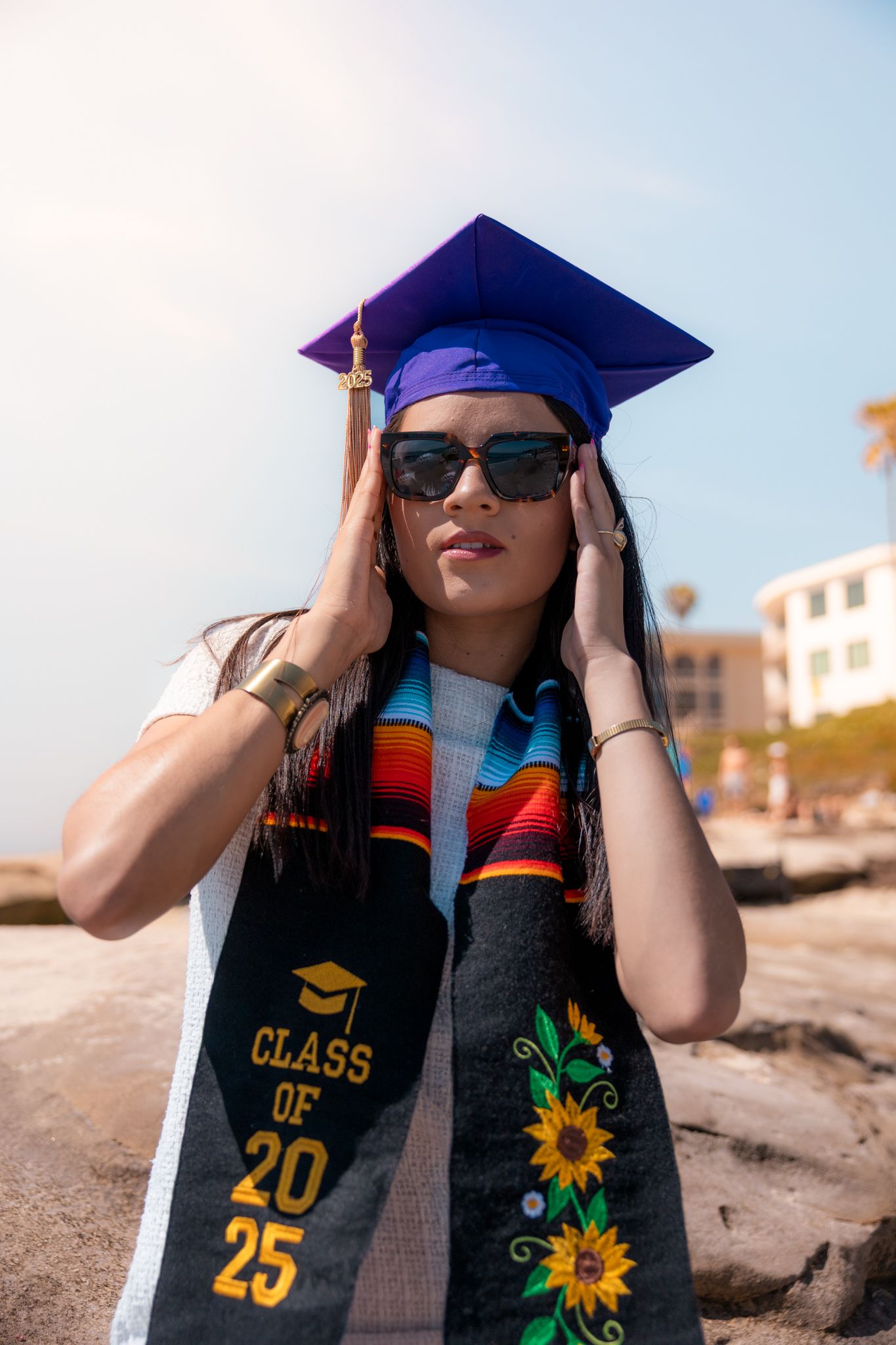 A graduate in a blue cap, sunglasses, and Class of 2025 stole poses on a rocky beach.