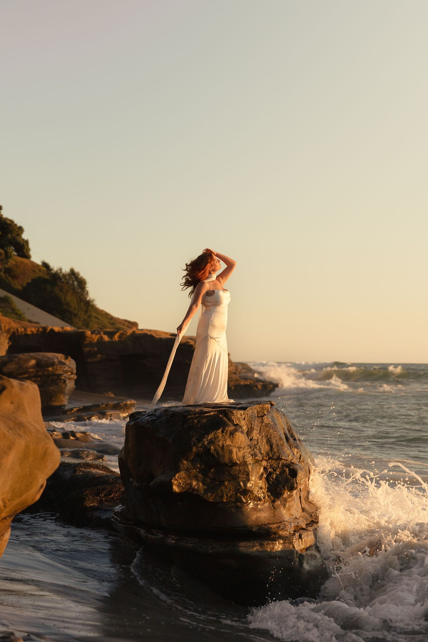 Woman in white dress standing on ocean rocks at sunset with waves crashing around her.