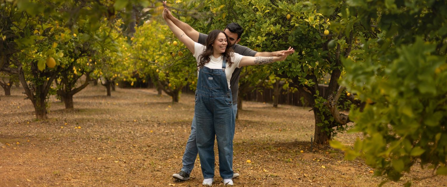 Couple in denim overalls posing with arms outstretched in a lush green orchard.
