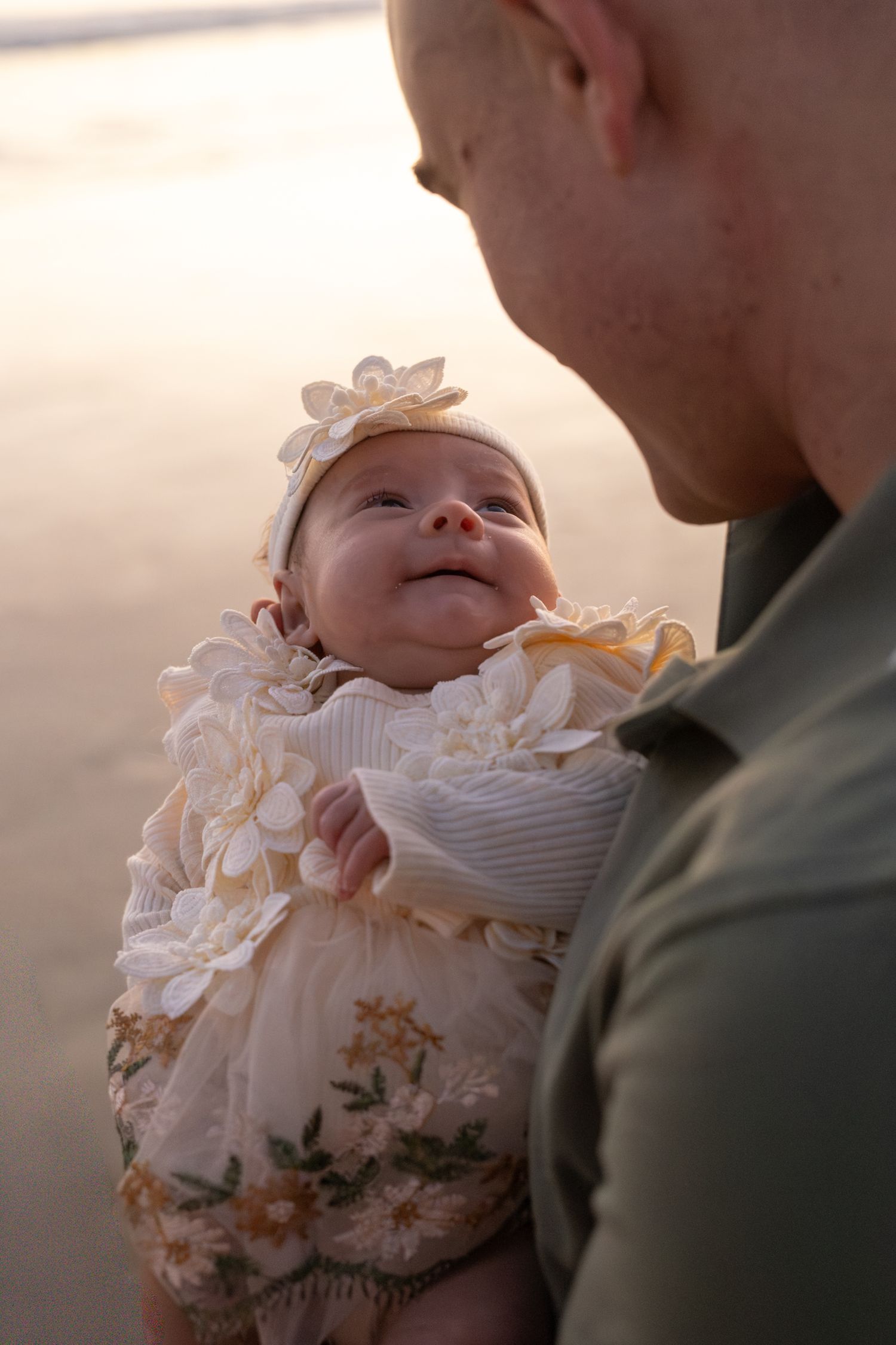 Bald father gazing lovingly at his smiling newborn baby dressed in a floral outfit on a beach.