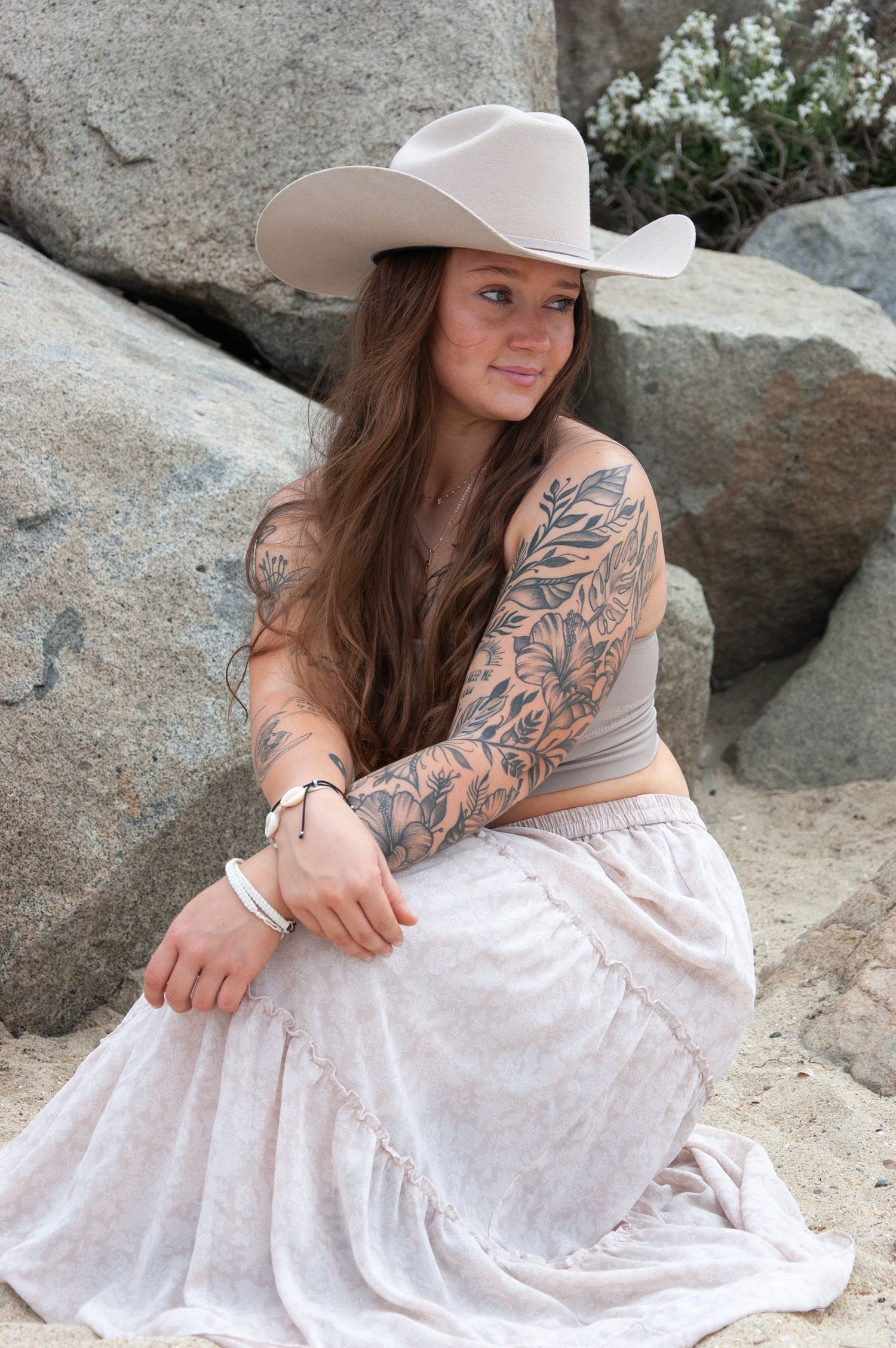 Young woman with sleeve tattoos wearing a cowboy hat and floral skirt near rocks.