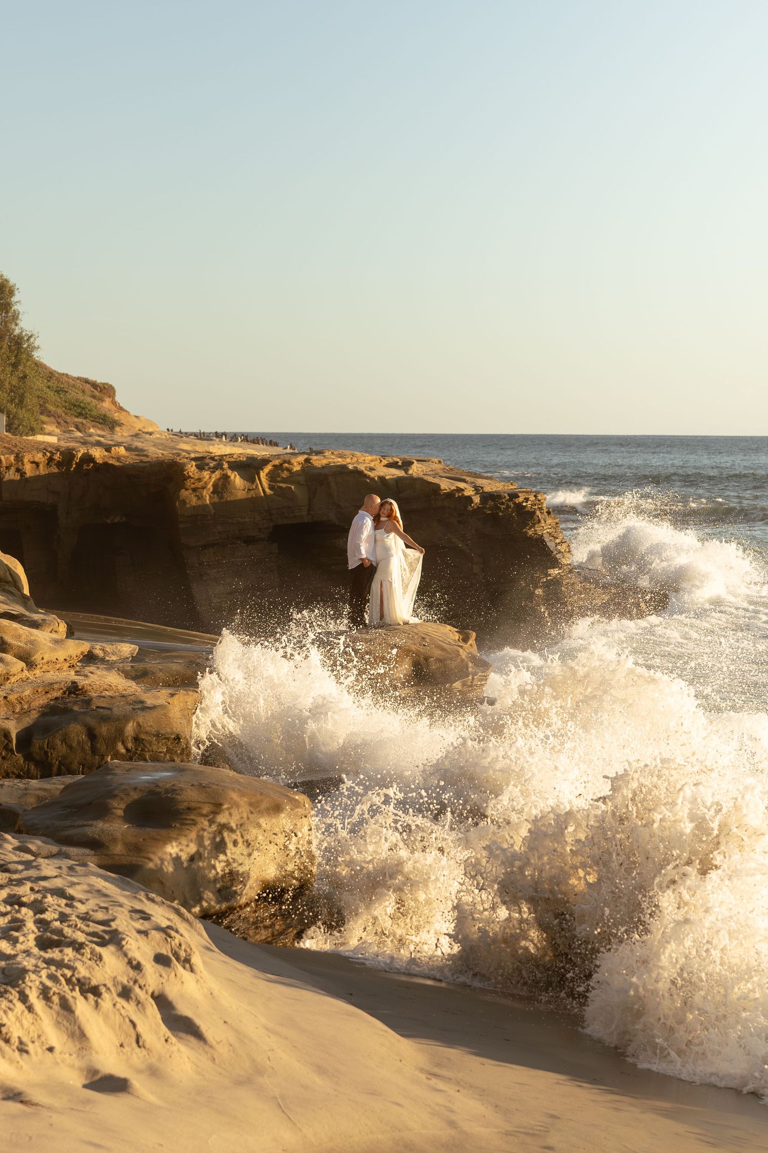 Bride and groom embrace on rocky shoreline as ocean waves crash around them at sunset.
