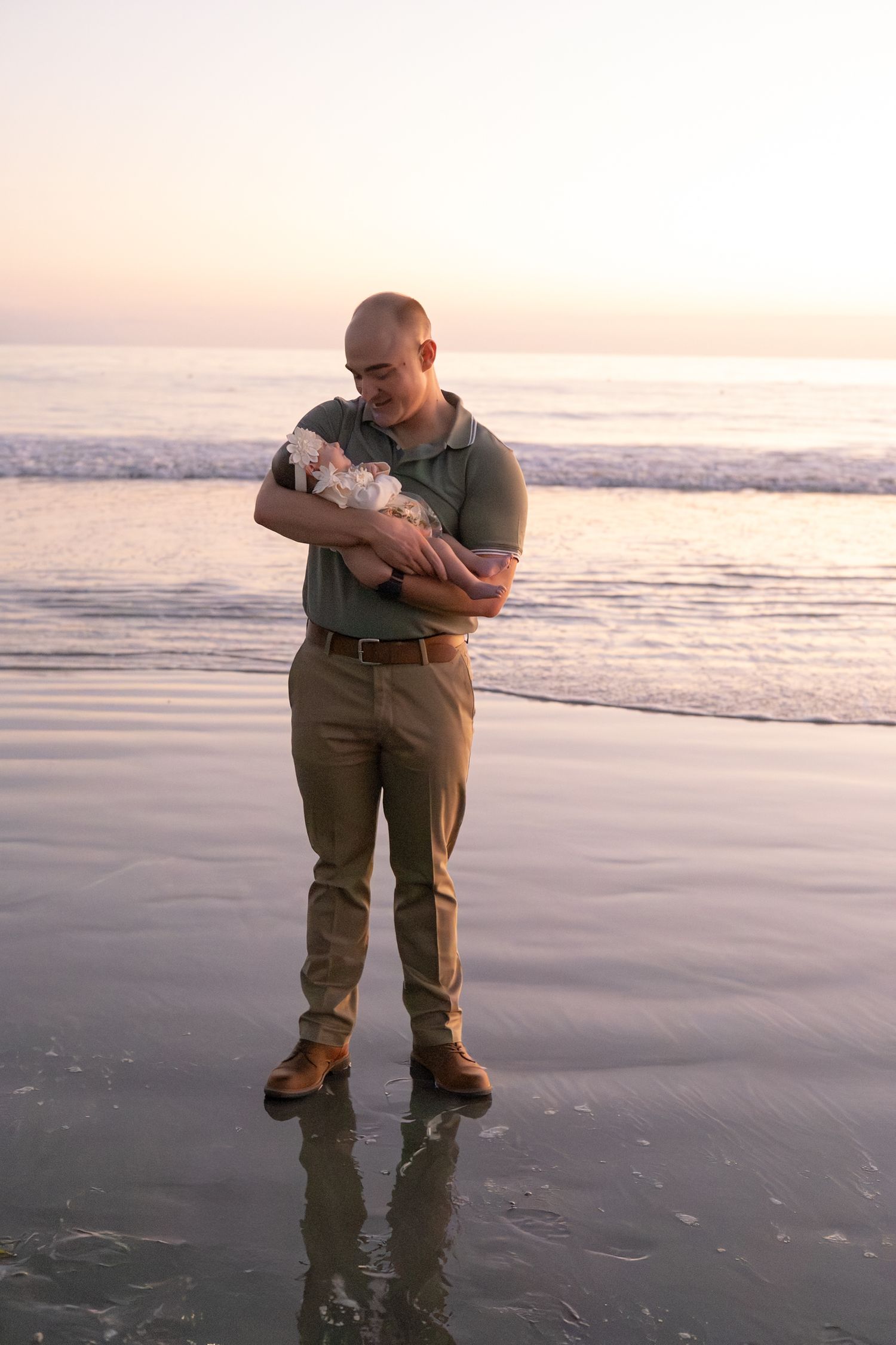 Bald father standing at ocean's edge at sunset while gently cradling his newborn baby.