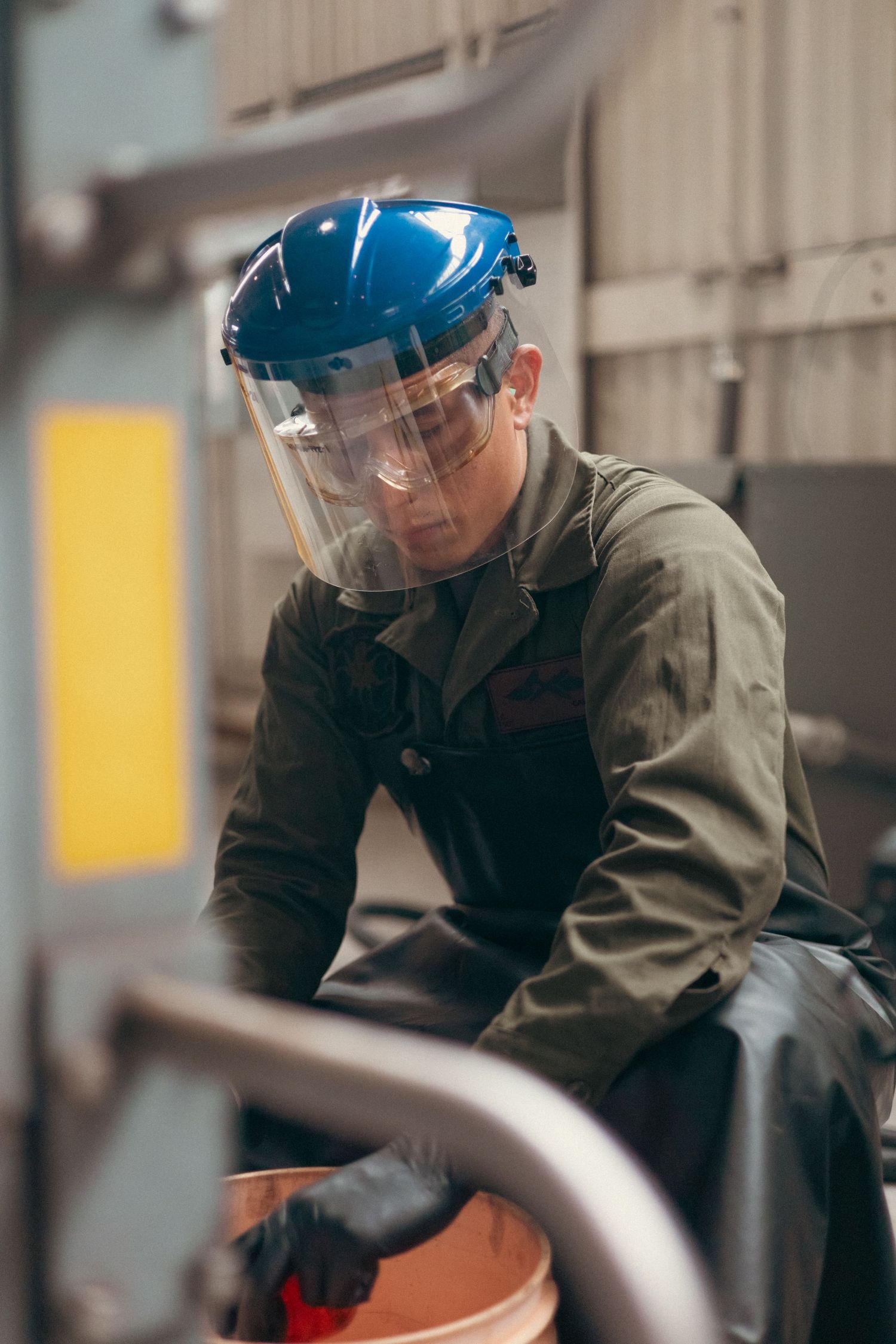 Industrial worker wearing blue hard hat and clear face shield handling equipment in workshop.