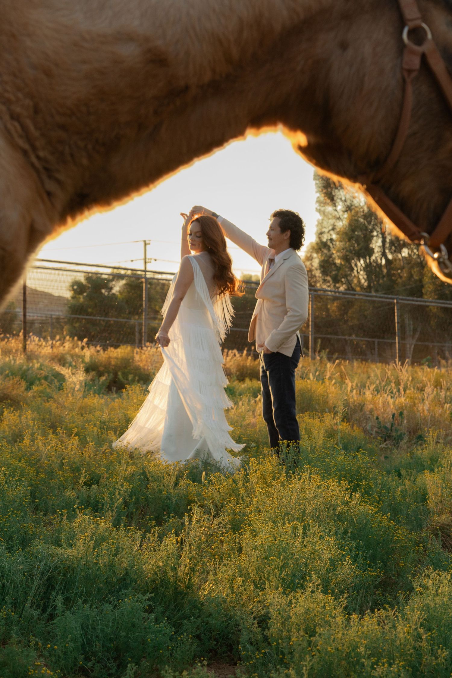 Bride and groom dancing in a golden sunset field, framed by a horse in a romantic outdoor wedding photo.