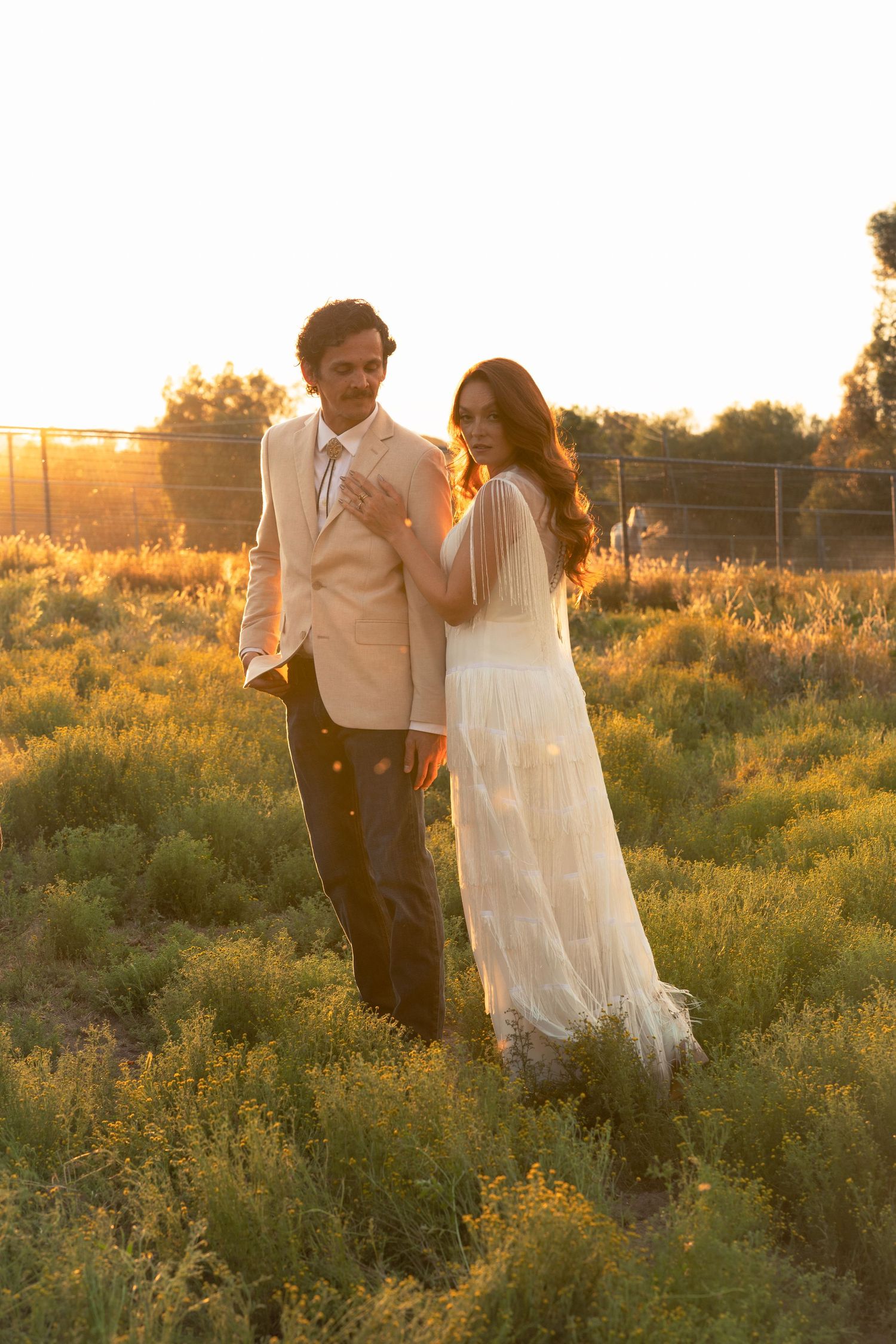 Couple in formal attire standing in golden field at sunset, woman in white gown, man in beige suit.