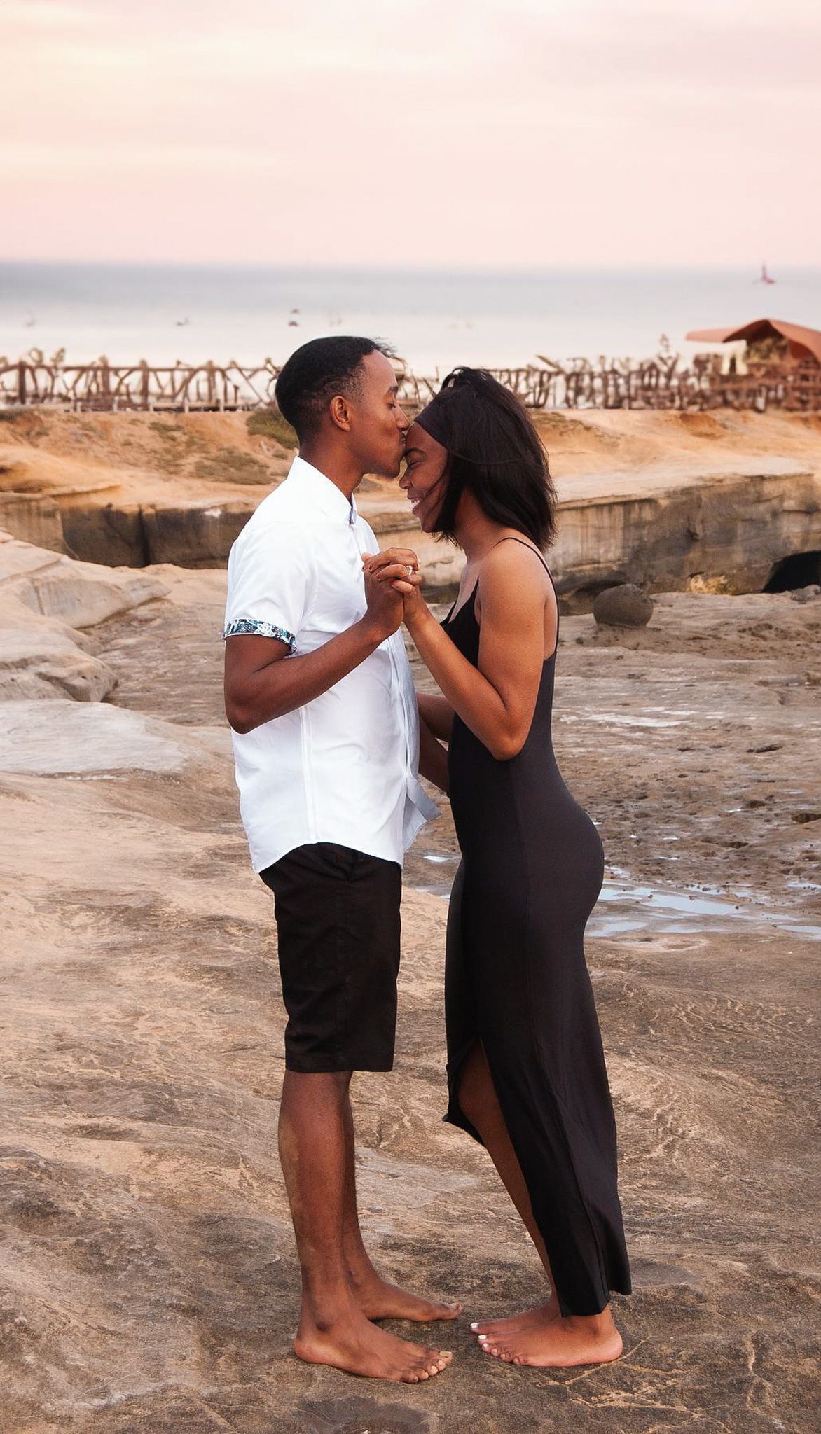 A couple shares a romantic moment on rocky coastal shores at sunset, holding hands.