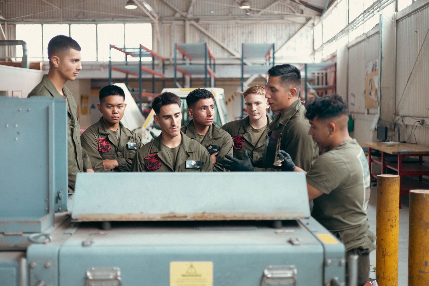 Group of Marines in olive uniforms examining military equipment inside a warehouse facility.