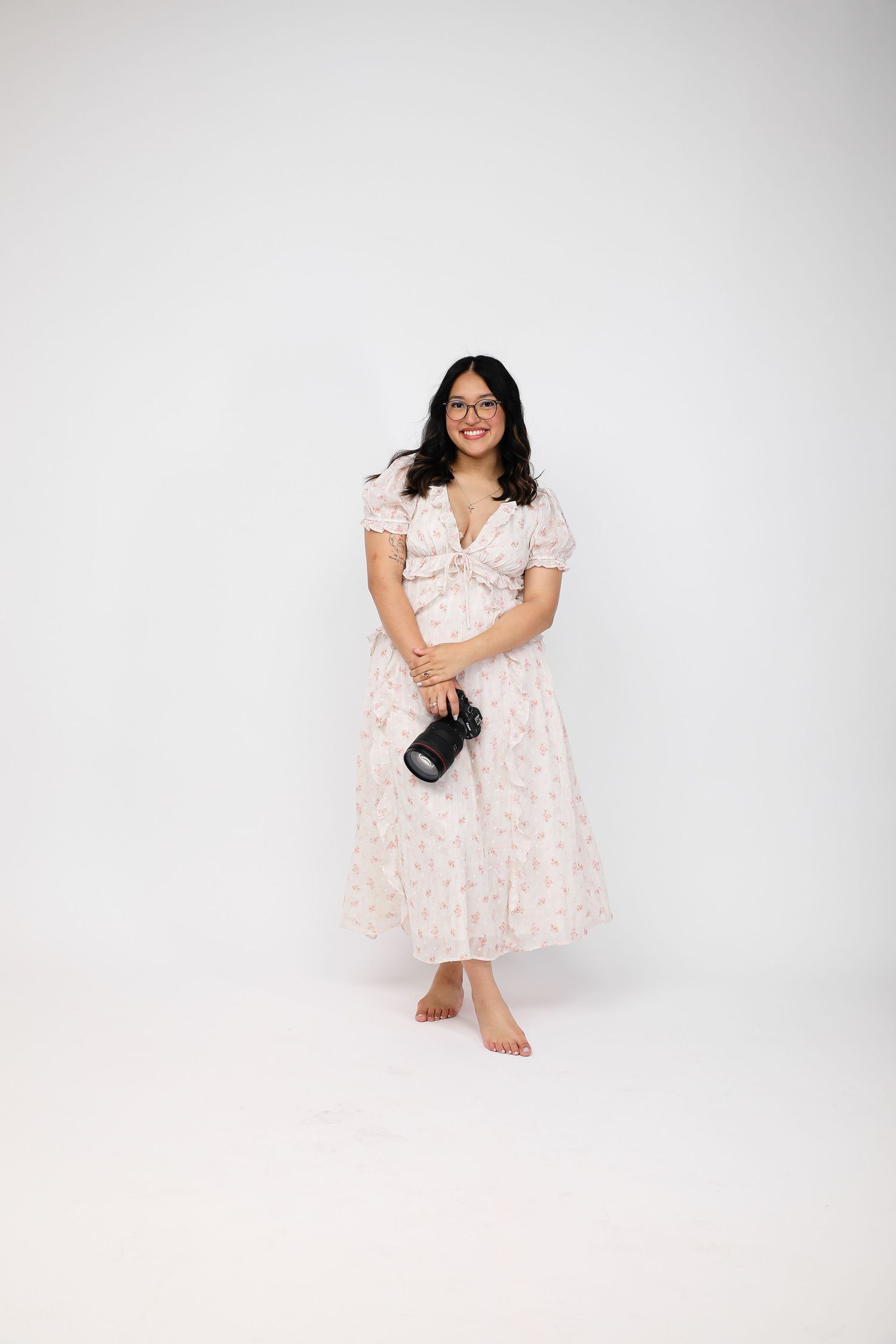Smiling woman in floral midi dress holding camera, standing barefoot against white background.