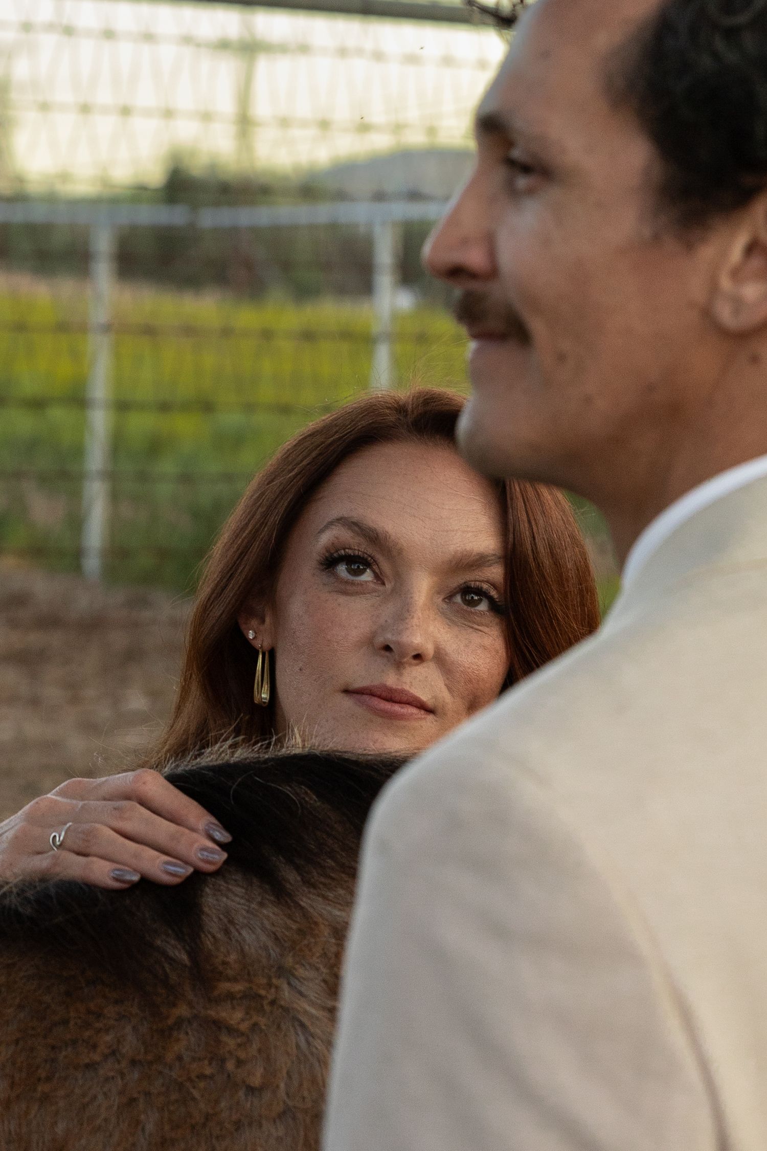 Redhead woman gazing at a man while petting a brown horse near a fence outdoors.