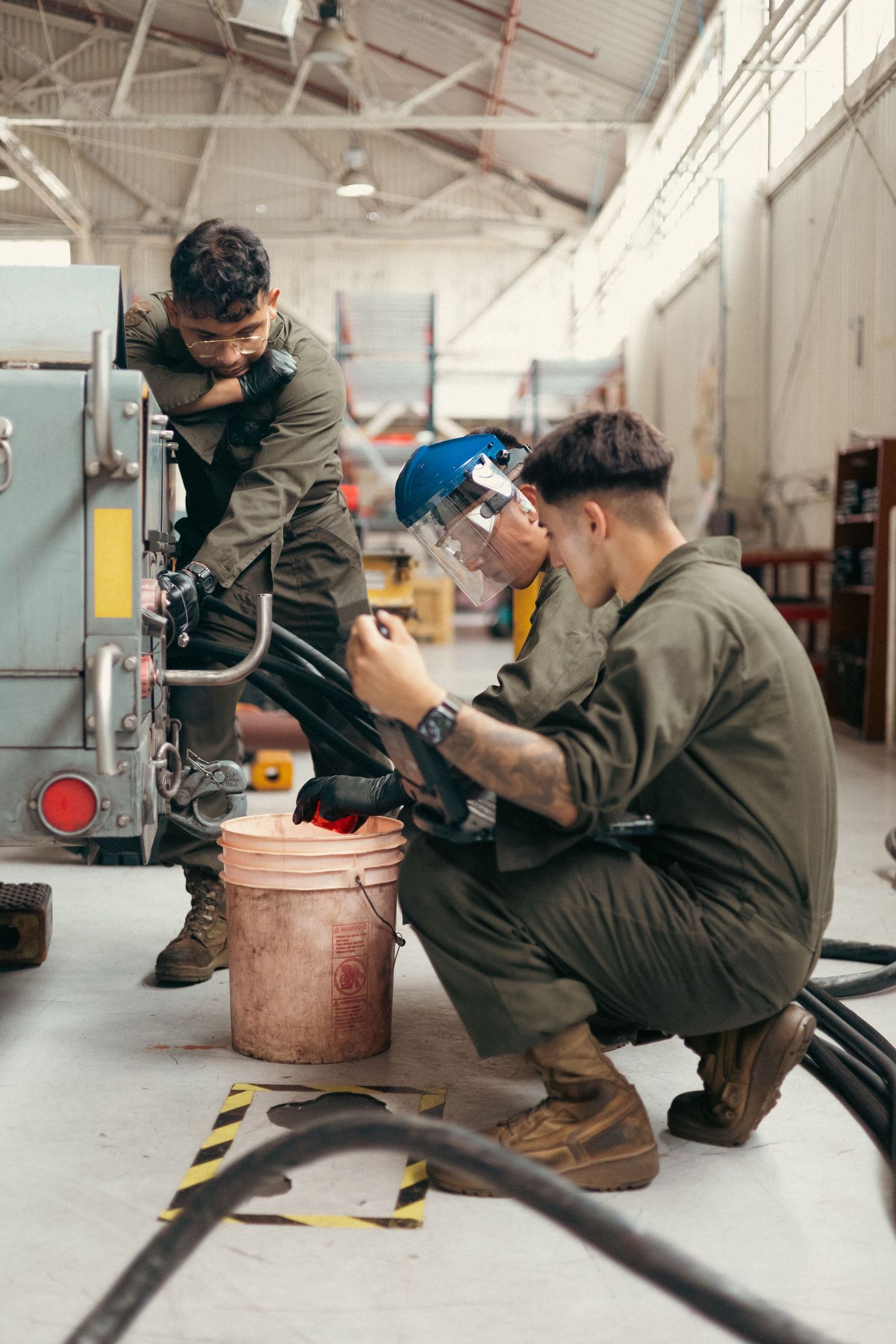 Three Marines in olive jumpsuits work together on industrial equipment in a warehouse using protective gear.