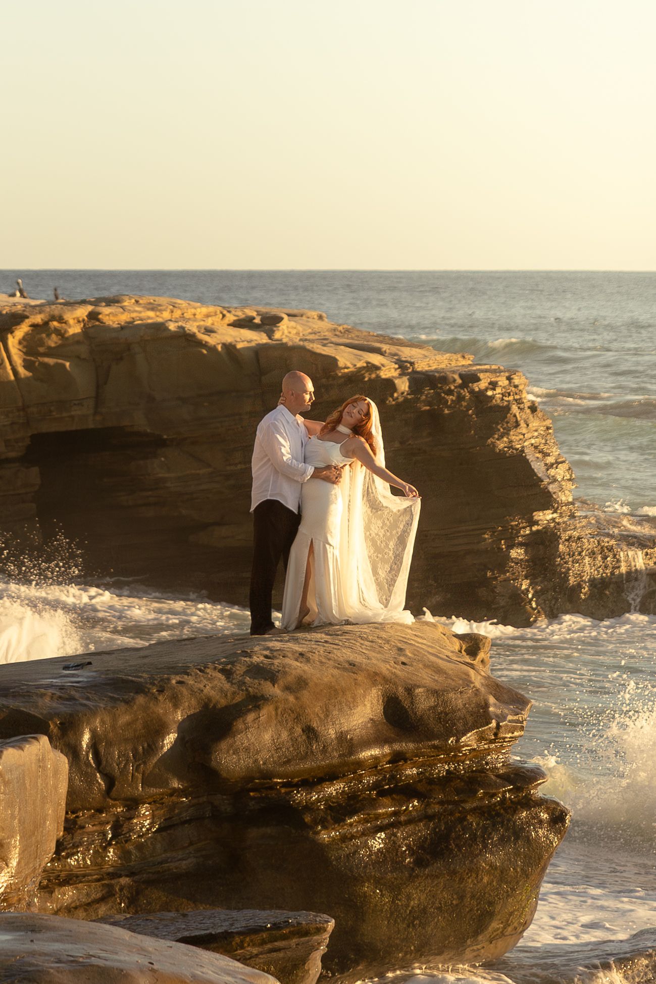 Couple in wedding attire embracing on rocky ocean cliffs at golden hour sunset by the sea.