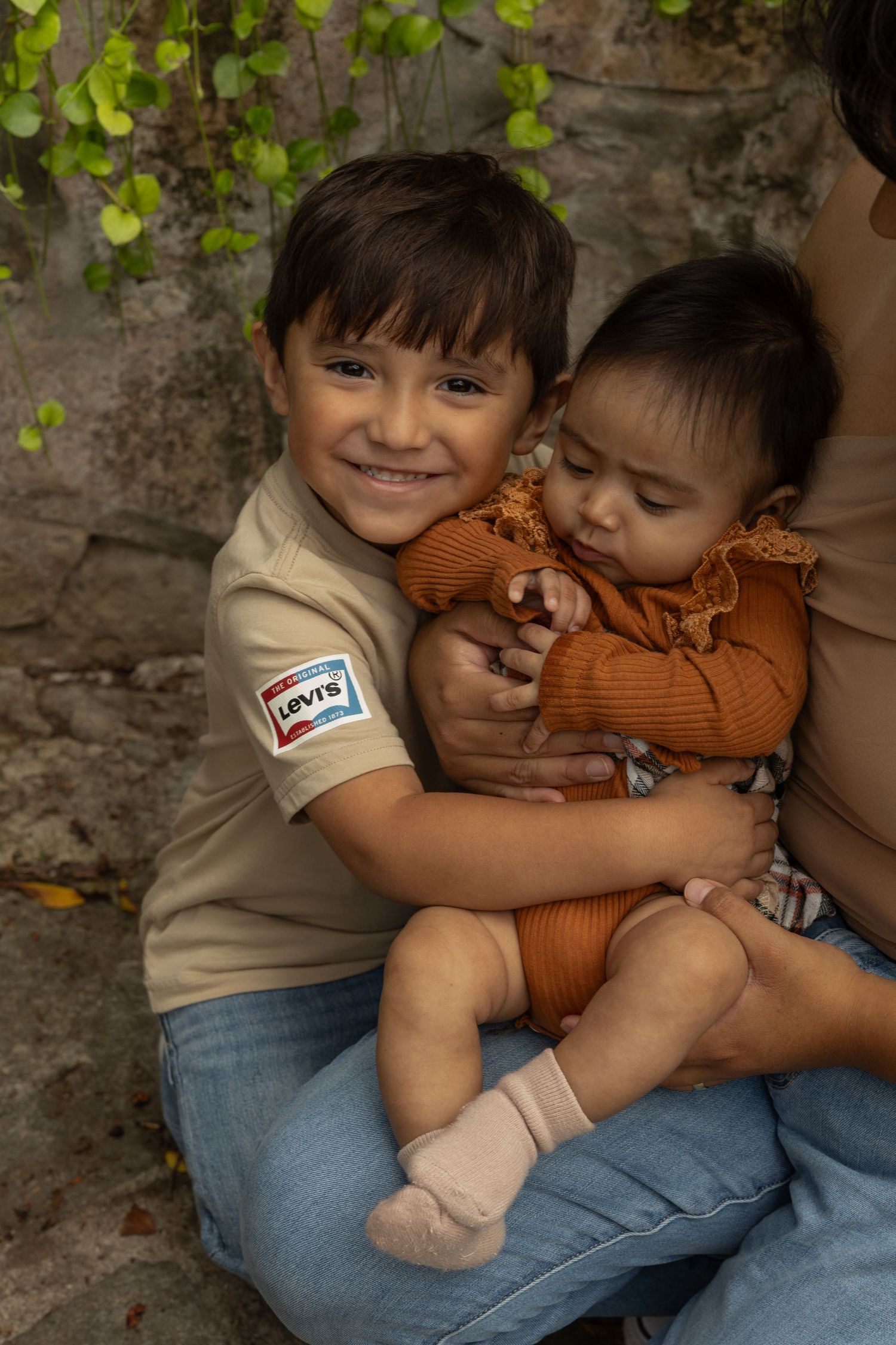 Smiling young boy in Levi's shirt tenderly holding a baby in an orange outfit outdoors.