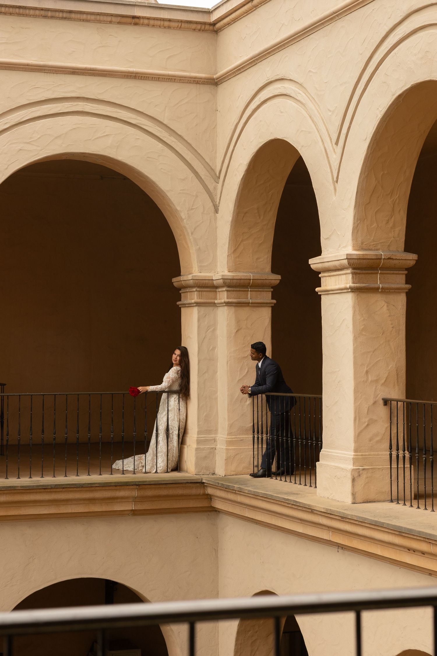 Bride and groom pose on an elegant arched balcony of a beige stone building during their wedding photo session.