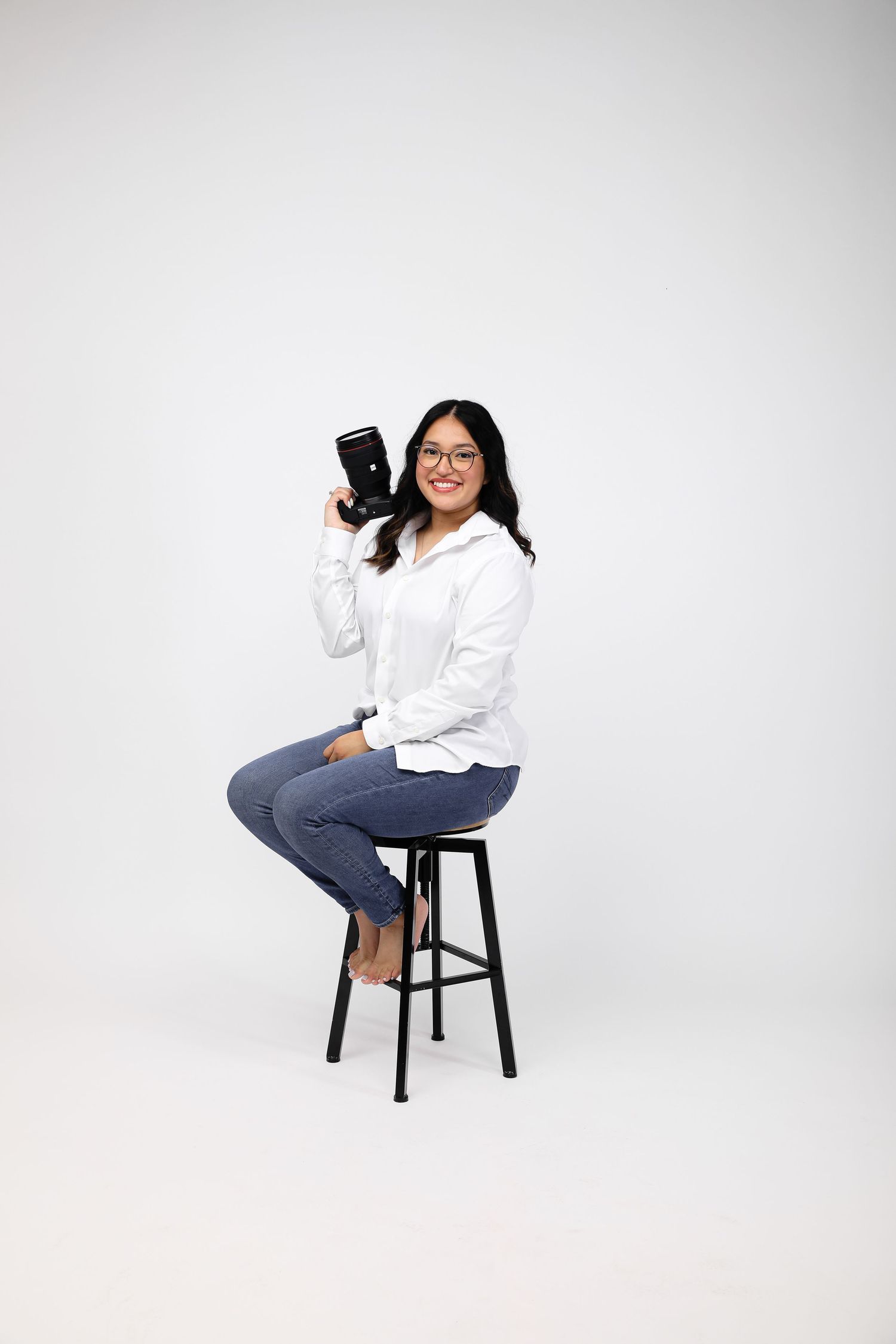 Woman in white shirt and jeans sitting on stool holding a camera, smiling against white background.