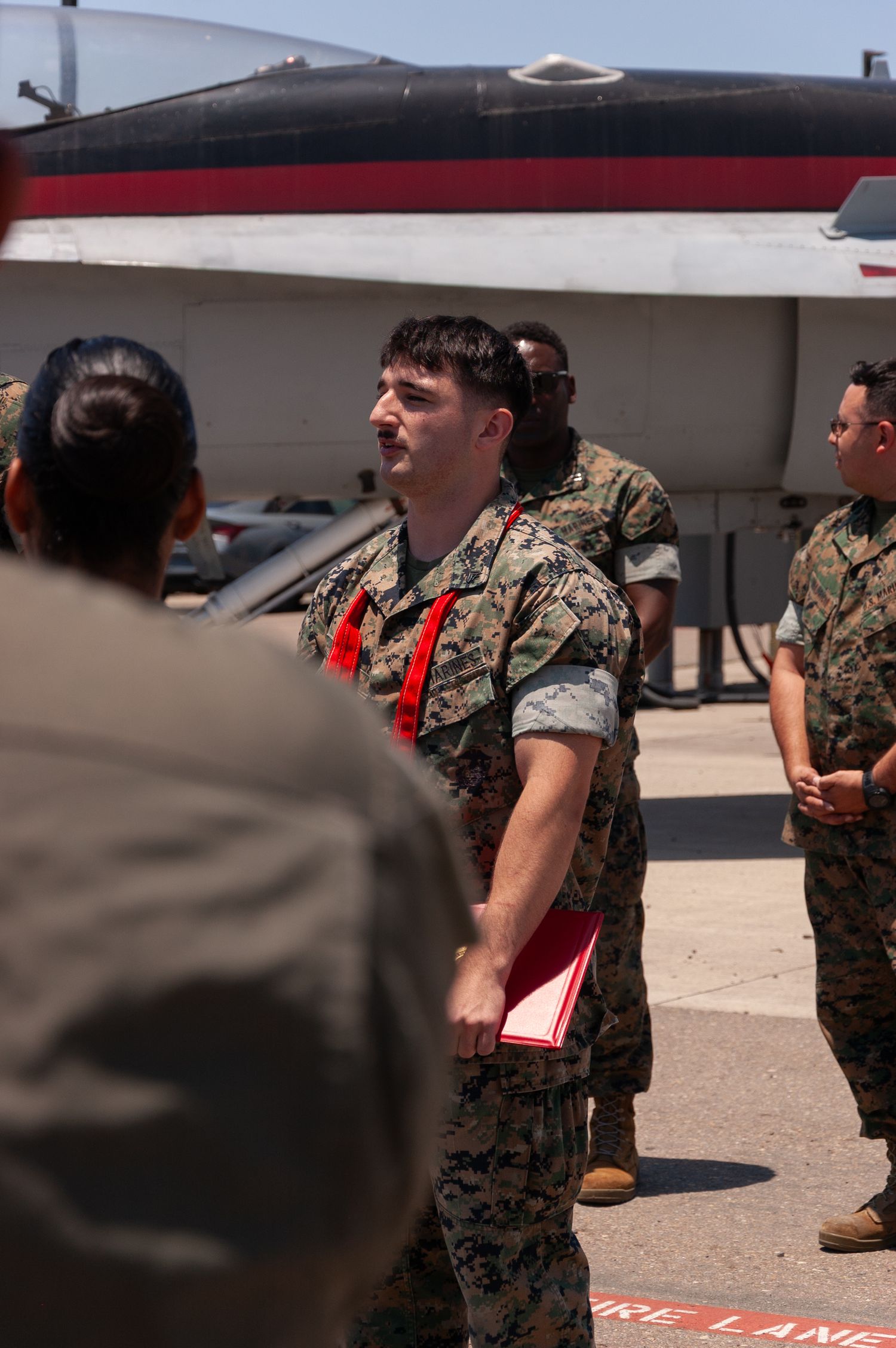 Marine in camouflage uniform addresses fellow Marines near red and white military aircraft.