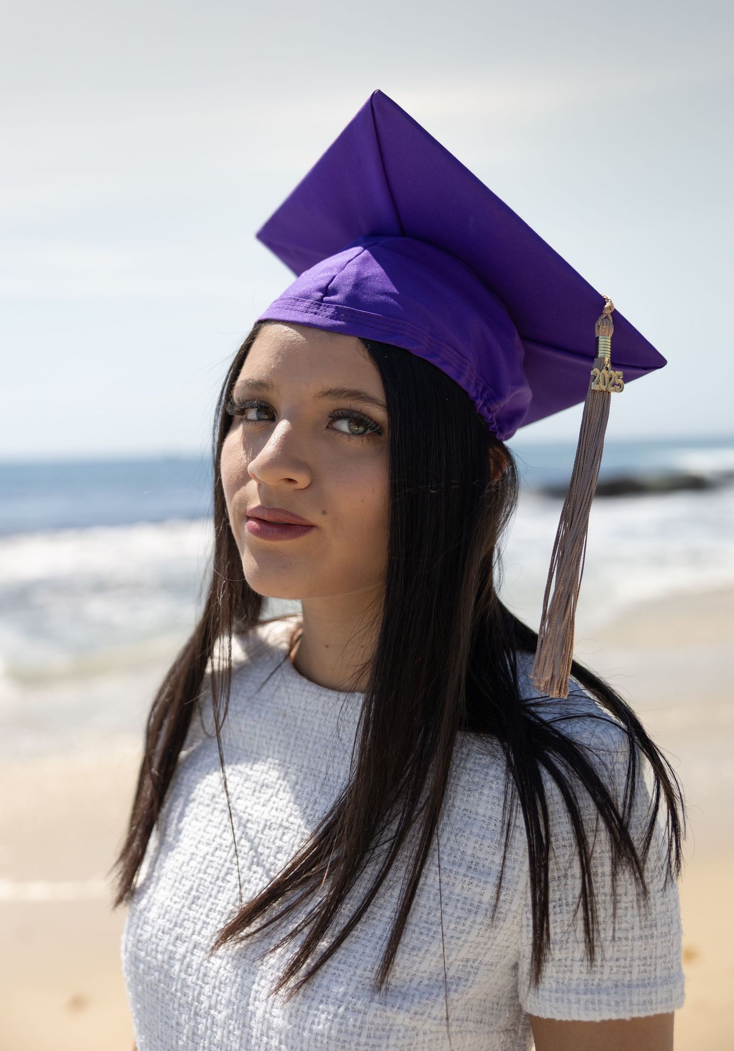 A young woman in a purple graduation cap with a tassel poses on a sunny beach.