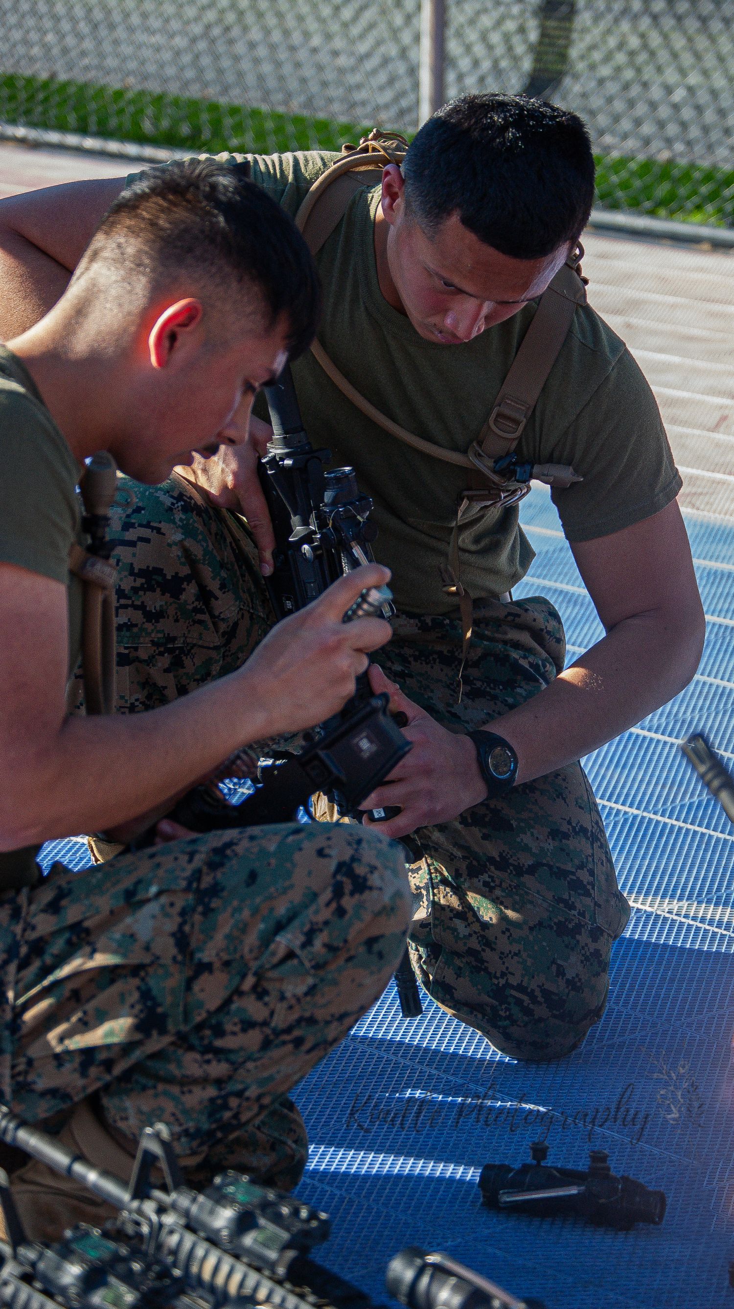 Two Marines in camouflage uniforms inspect equipment while kneeling on blue mat outdoors.