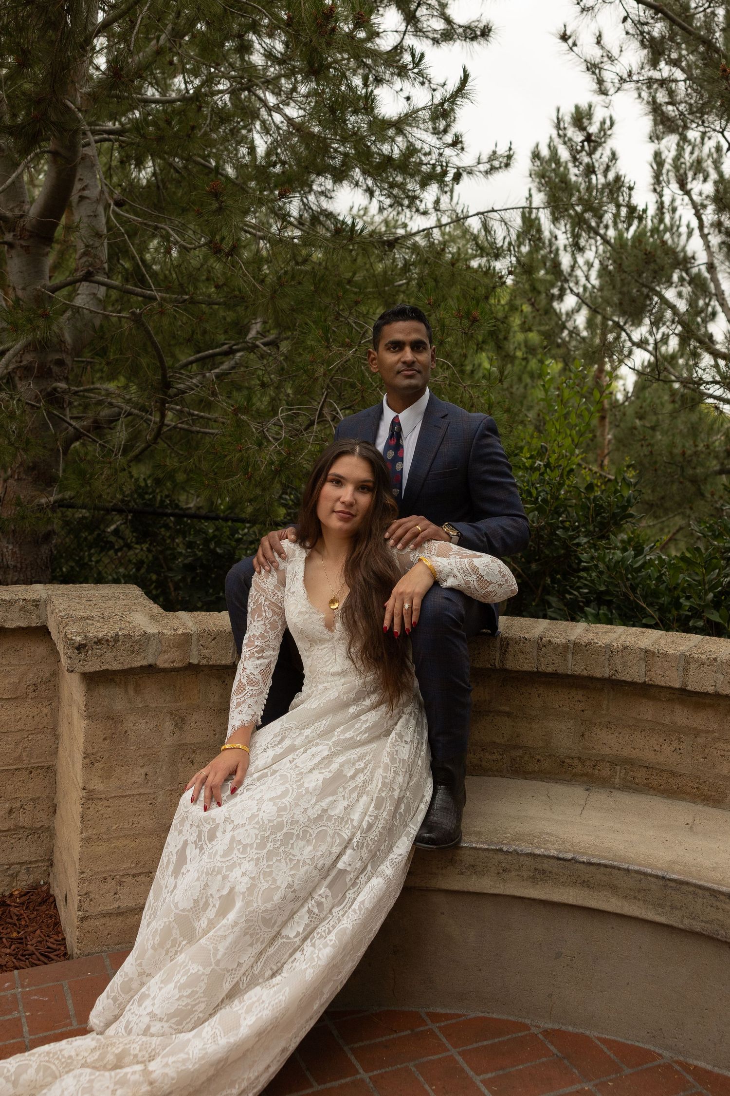 Bride in white lace gown and groom in navy suit pose together at an outdoor stone venue surrounded by trees.