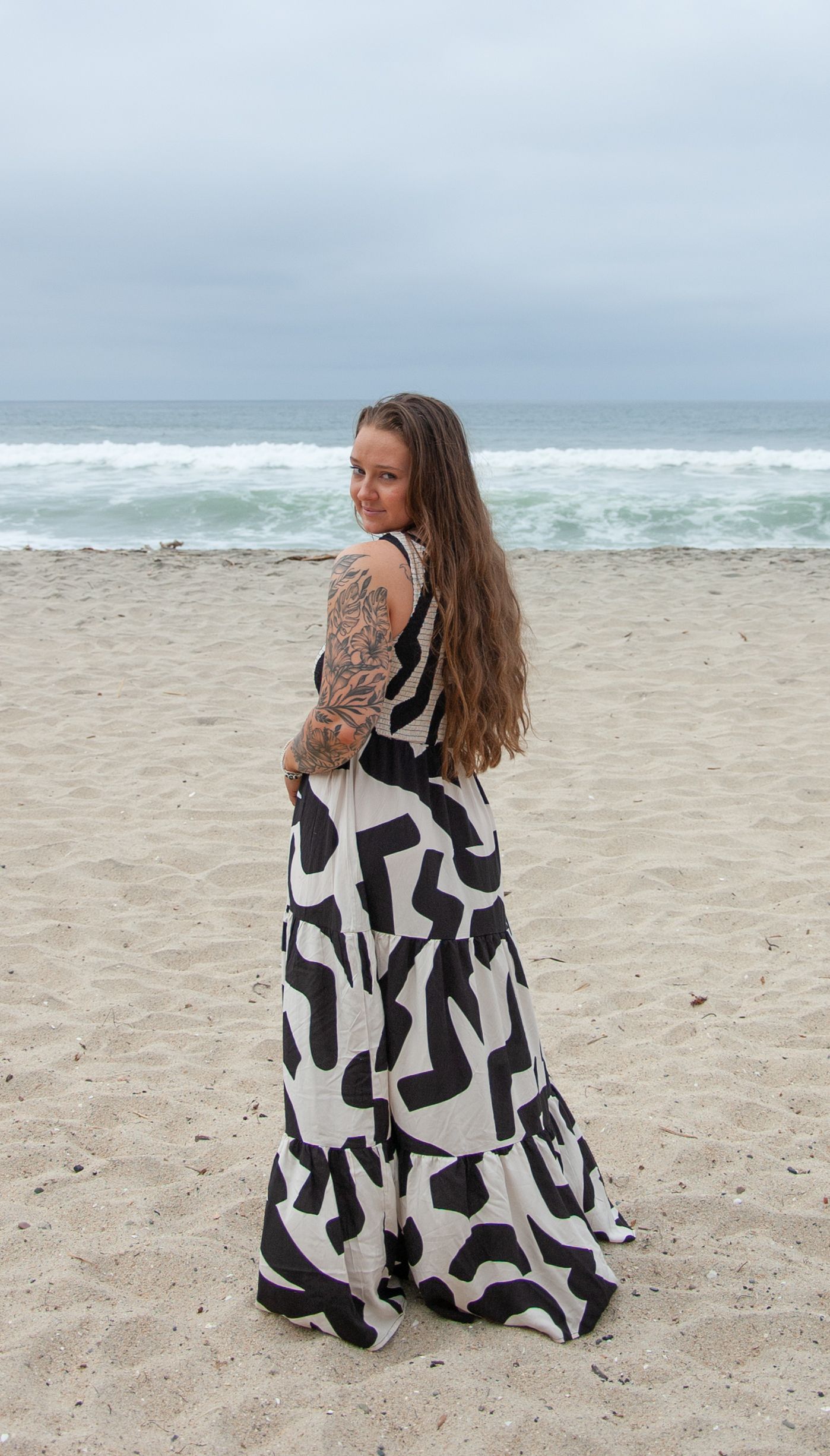 Tattooed woman in black and white maxi dress standing on a beach looking back.