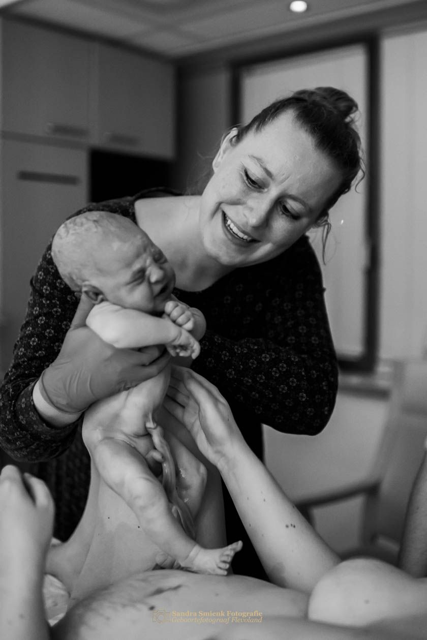 Black and white photo of a smiling woman holding a newborn baby moments after birth in a hospital room. Lelystad, Almere, Dronten, Flevoland
