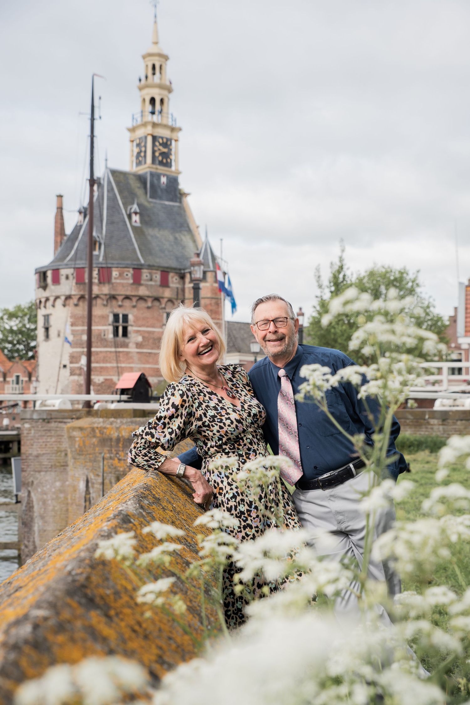 Smiling couple posing by a stone bridge with white wildflowers and a historic Dutch clock tower in the background.
