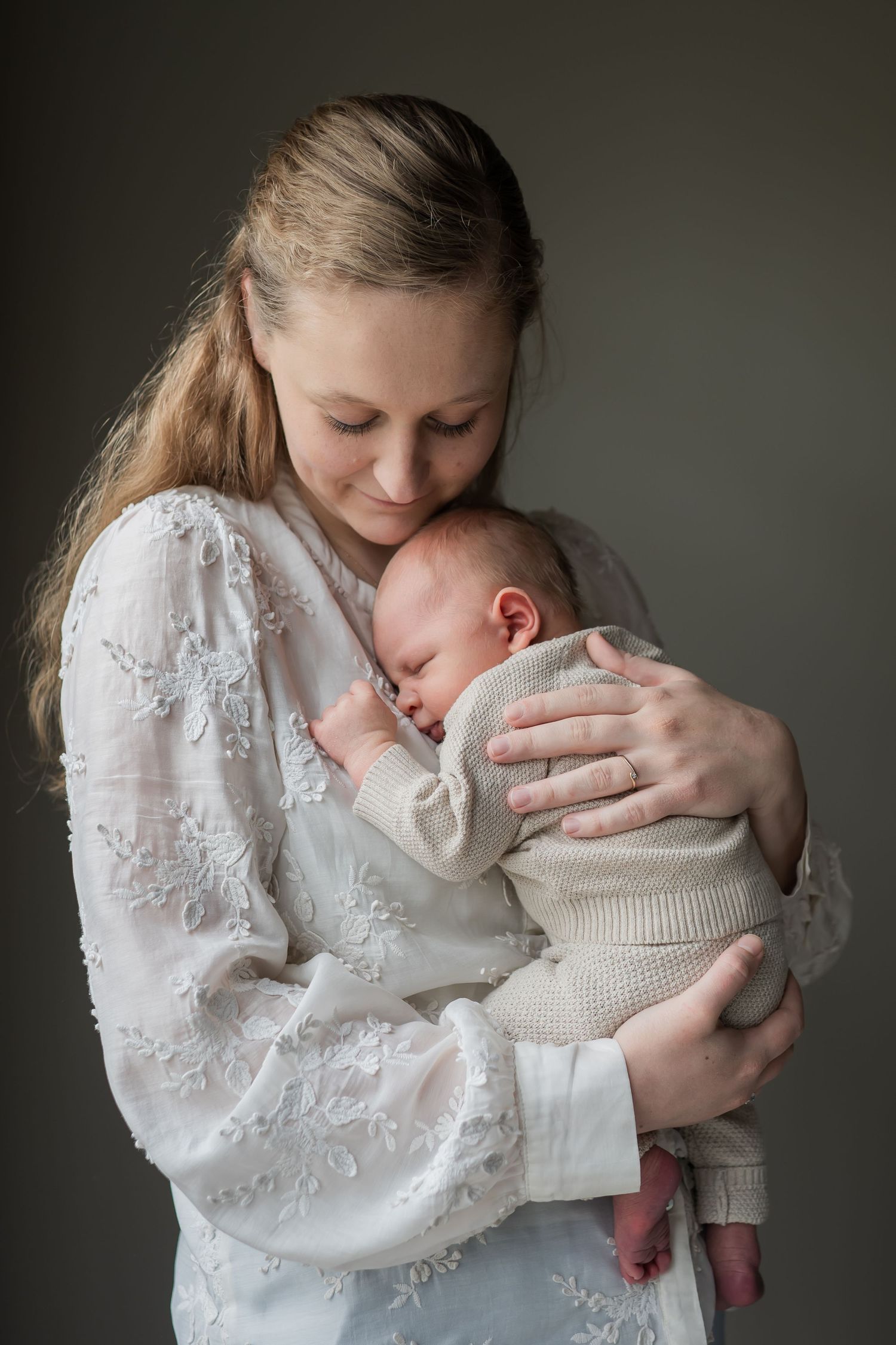 A mother in a white lace dress tenderly holds her sleeping newborn baby close to her chest in a studio portrait.