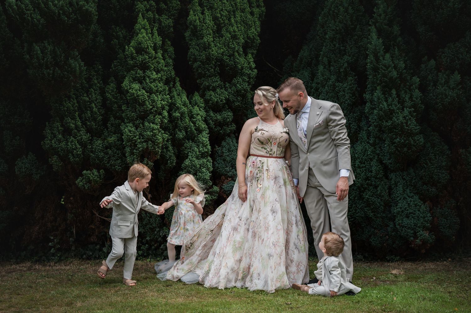 Wedding couple in floral gown and tan suit pose with three young children on green lawn near tall evergreen trees.