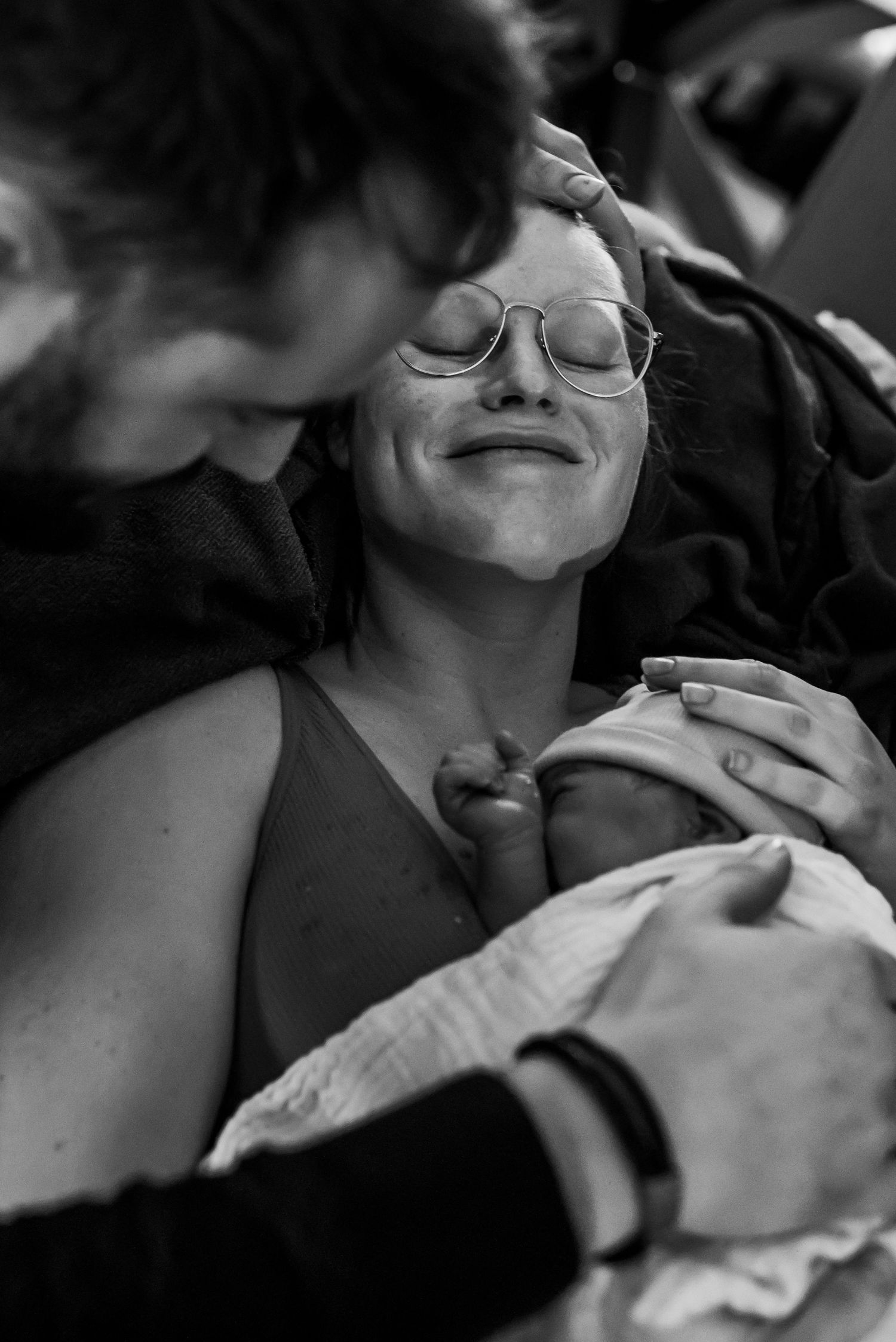 Black and white photo of a smiling new mother holding her newborn baby, supported by a partner stroking her hair. Lelystad, Almere, Dronten,