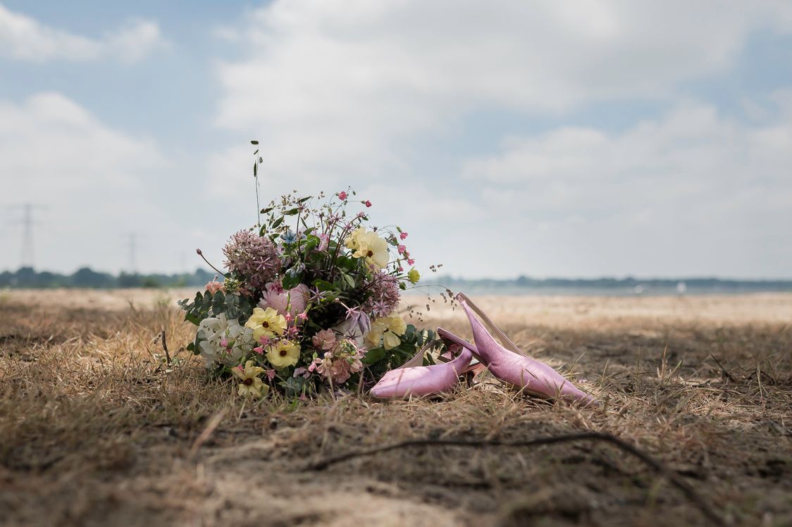 Colorful wildflower wedding bouquet with pink ribbons lying on dry grass field under cloudy sky.