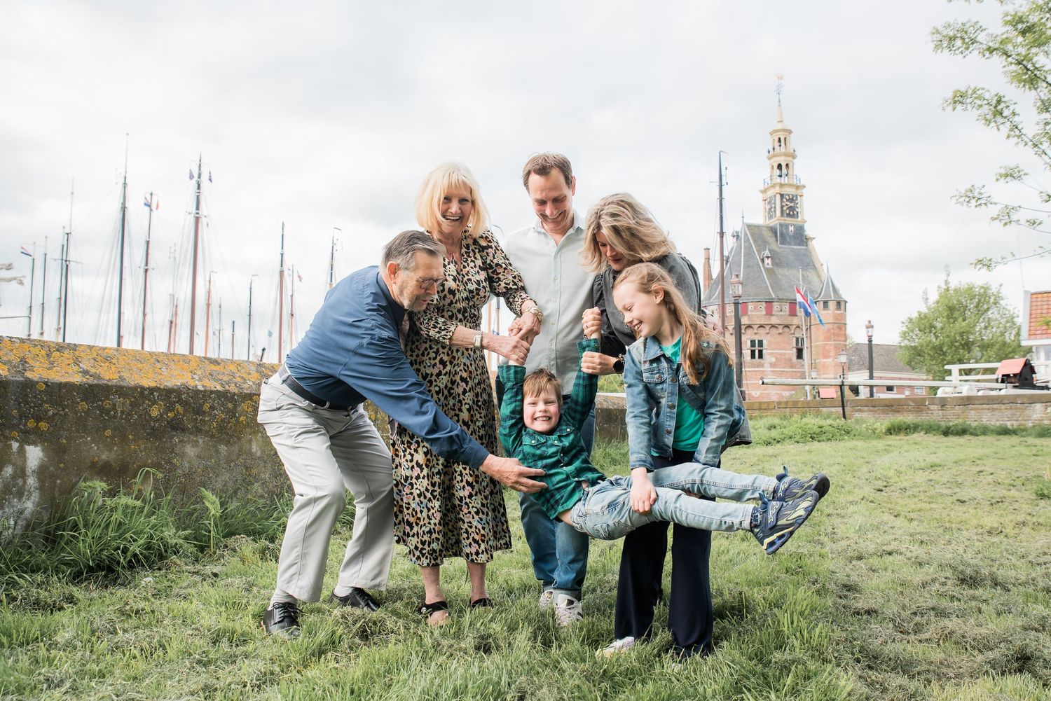 Multi-generational family laughing and posing outdoors on grass near historic Dutch building on cloudy day.