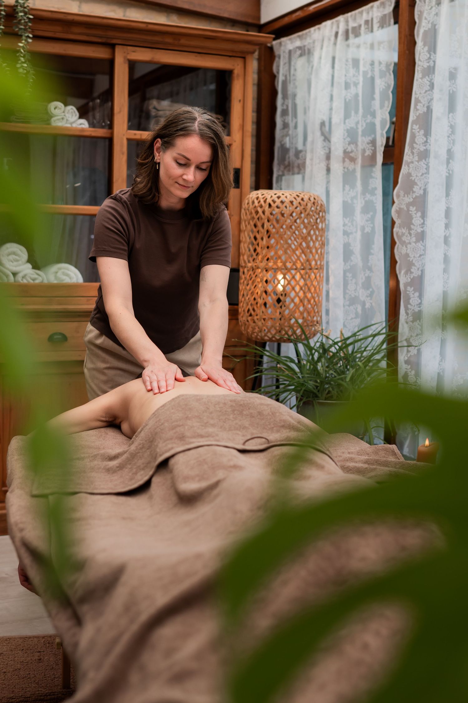 A female massage therapist in brown uniform performs a relaxing back massage on a client in a cozy spa room.