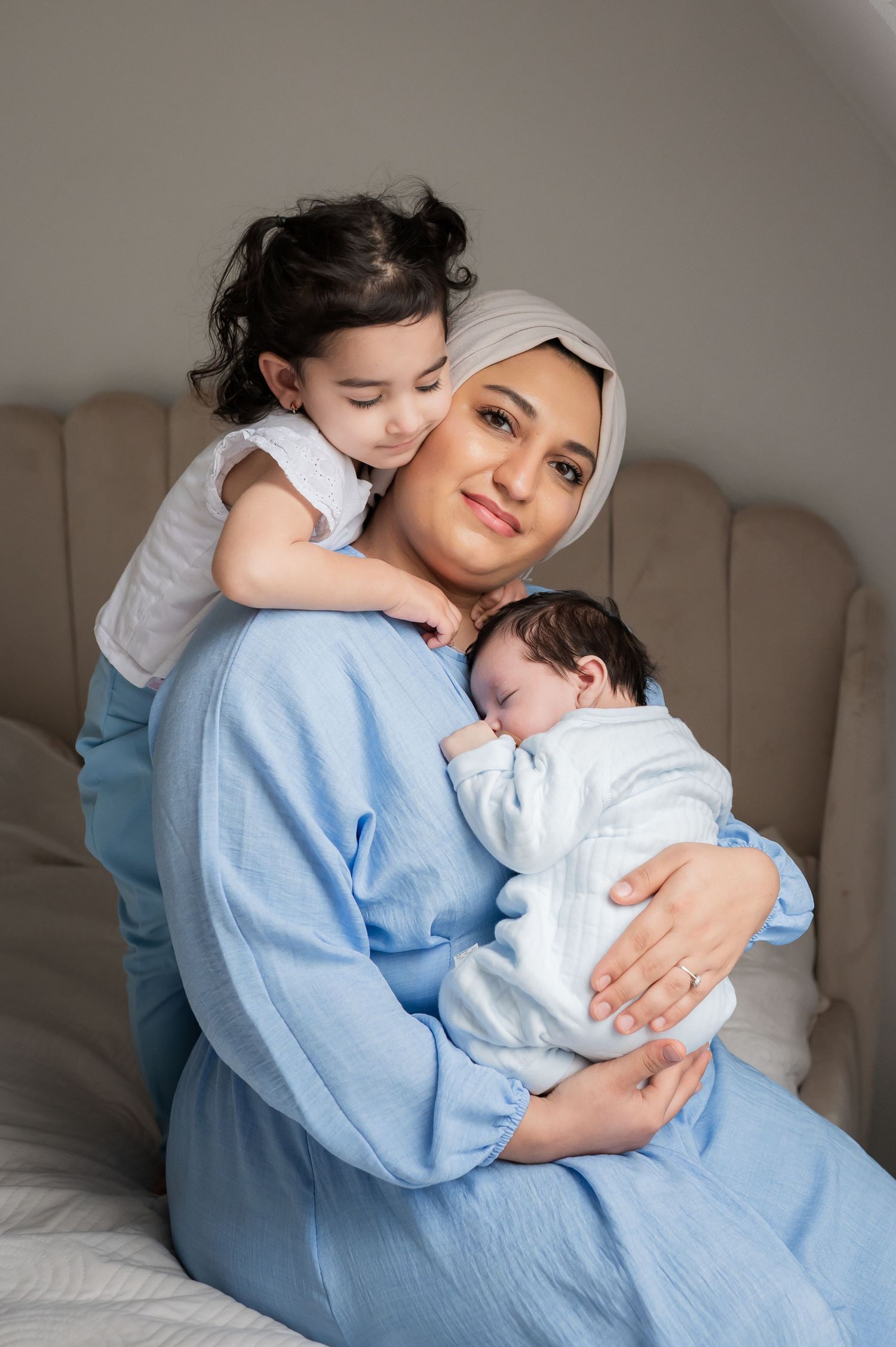 Smiling mother in blue dress wearing hijab holding newborn while toddler daughter hugs her from behind.