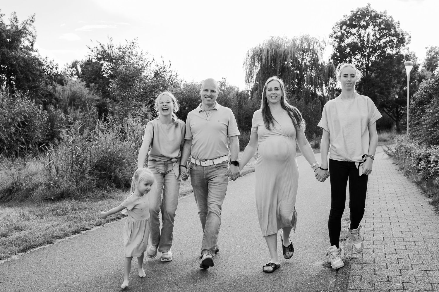 Black and white photo of a pregnant woman walking with her family on a park path, holding hands outdoors.